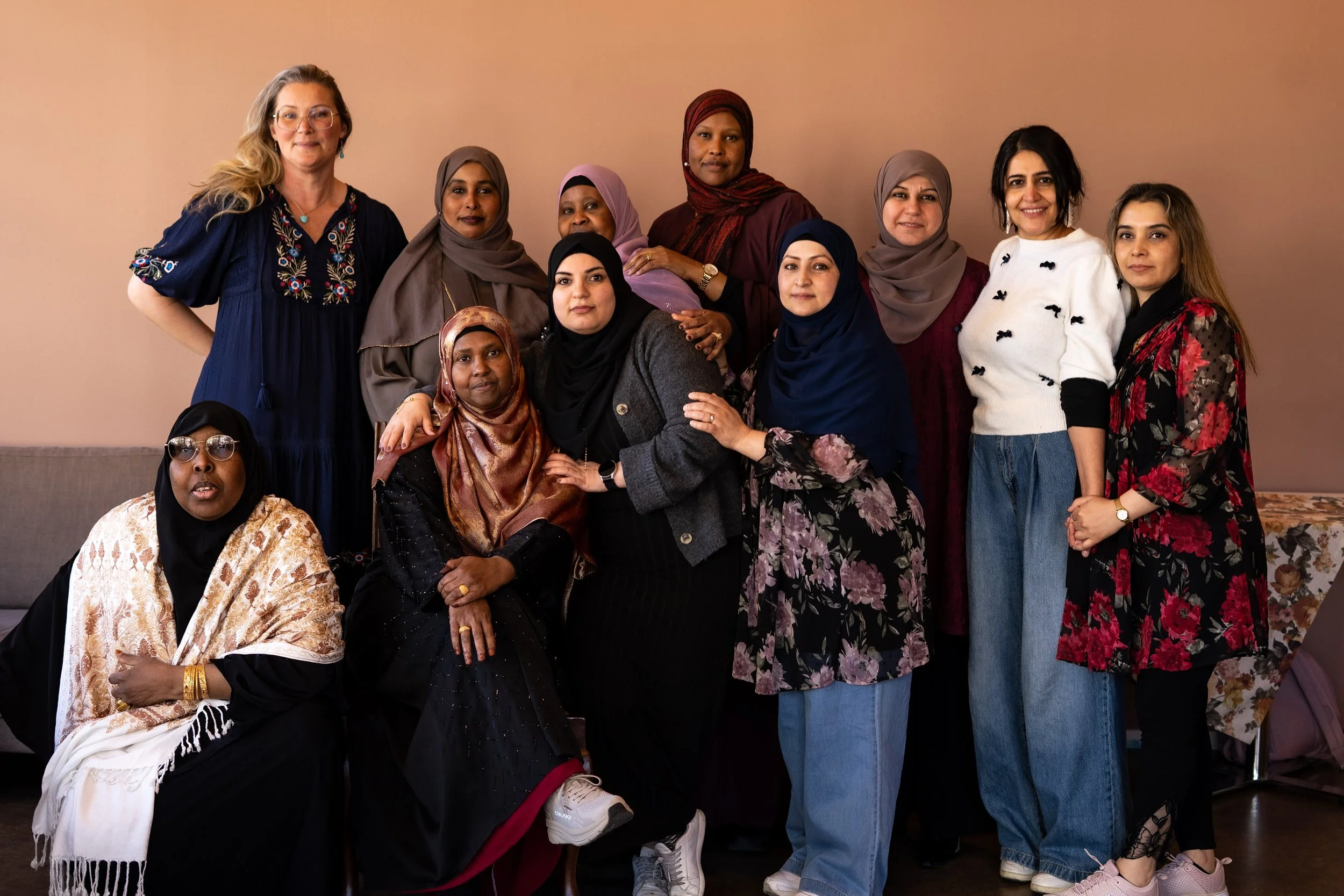 En grupp kvinnor står tillsammans inomhus framför en beige vägg, de flesta är iklädda hijabs och moderna kläder, och några är leende medan andra ser allvarliga ut.