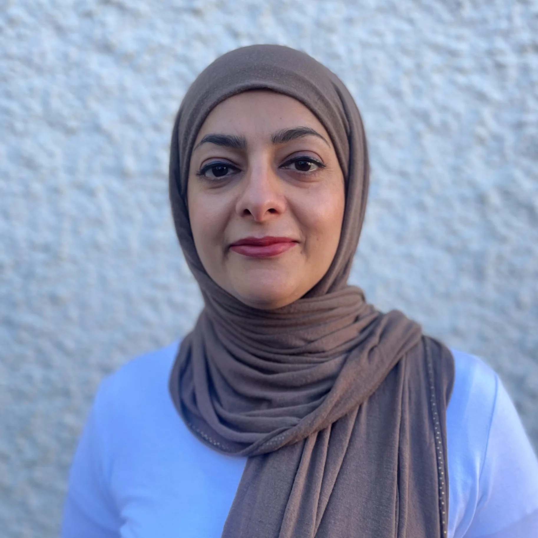 En kvinna bär en ljusbrun hijab och röd läppstift, tar en selfie mot en ljus, grynig bakgrund.