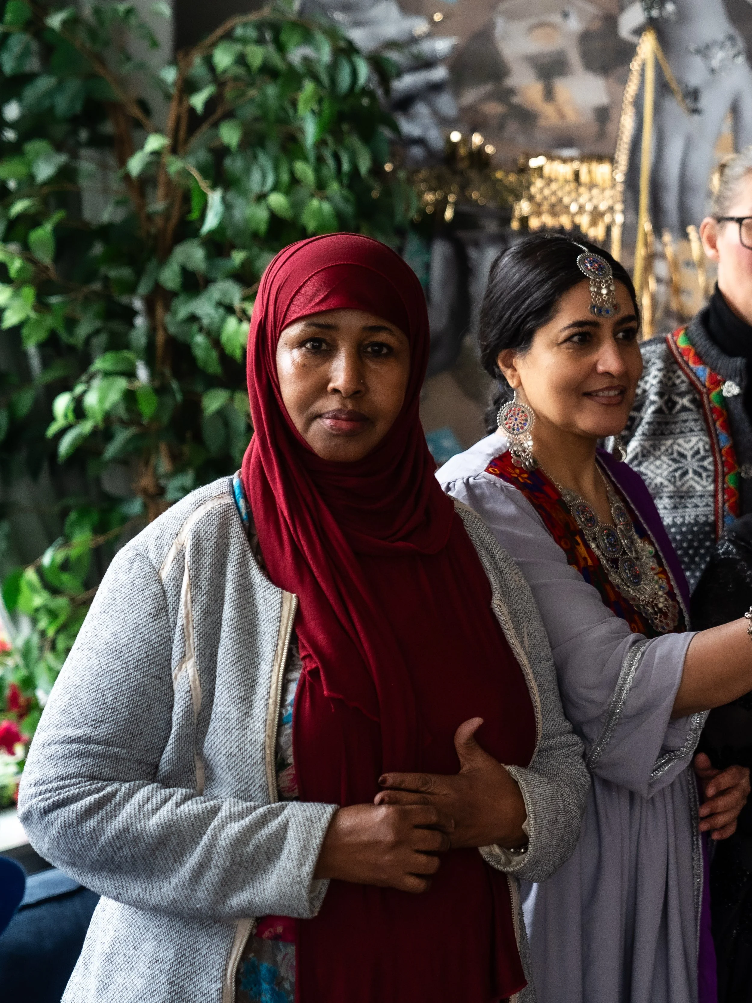 Tre kvinnor som står nära varandra, den första kvinnan bär en röd slöja och en grå jacka, den andra kvinnan bär traditionella smycken och ett mönstrat plagg, den tredje personen syns delvis till höger och är utomhus.
