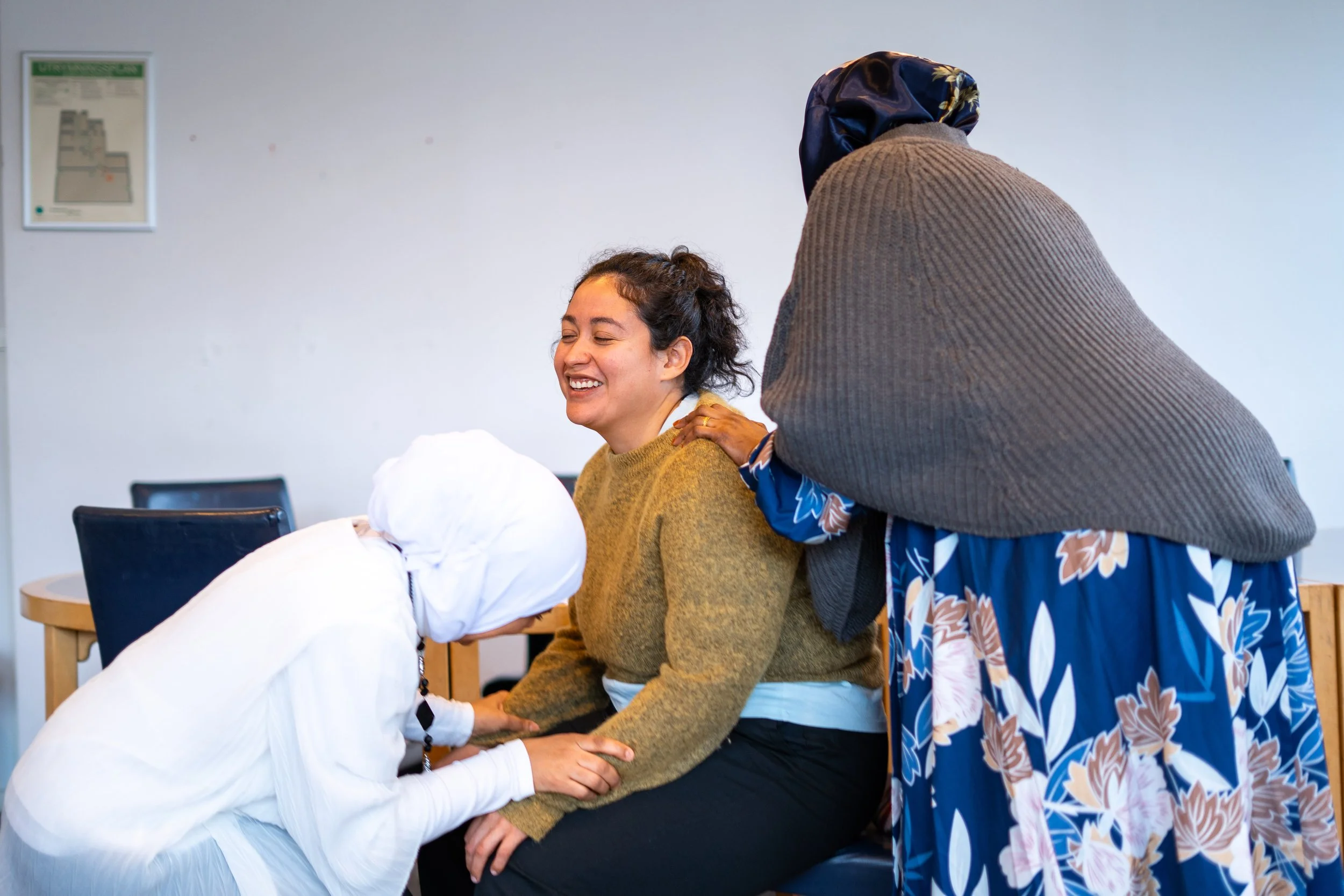 Tre personer i ett mötesrum som ler och håller varandra i händerna, inklusive en person i vit arbetsdress, en kvinna i en brungul tröja och en kvinna i en blå klänning med blommönster och ett grått huvudbonad.
