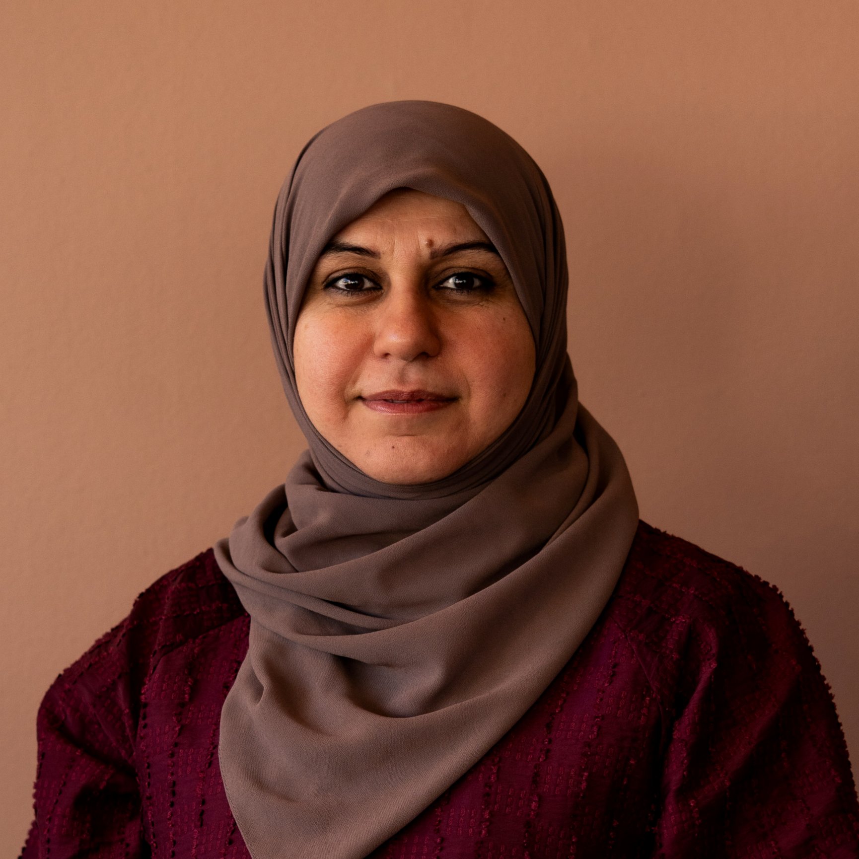 Porträtt av en kvinna med hijab i beige till en lätt rosa bakgrund, hon har ljust leende och mörka ögon.