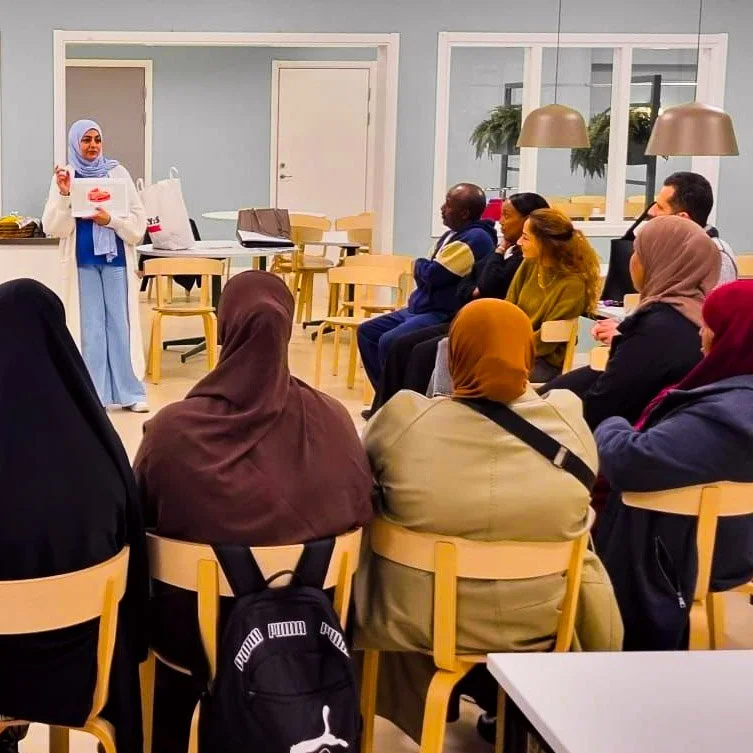 En kvinna som håller ett föredrag för en grupp människor i ett modernt rum. Alla är sittande och lyssnar, vissa med huvudet täckta av hijab.