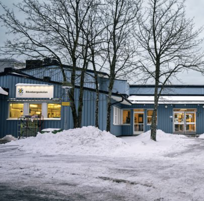En blå byggnad med ett skylt som säger 'Blåbergaskolan', omgiven av snö och tre träd utan löv, på vinterkväll.