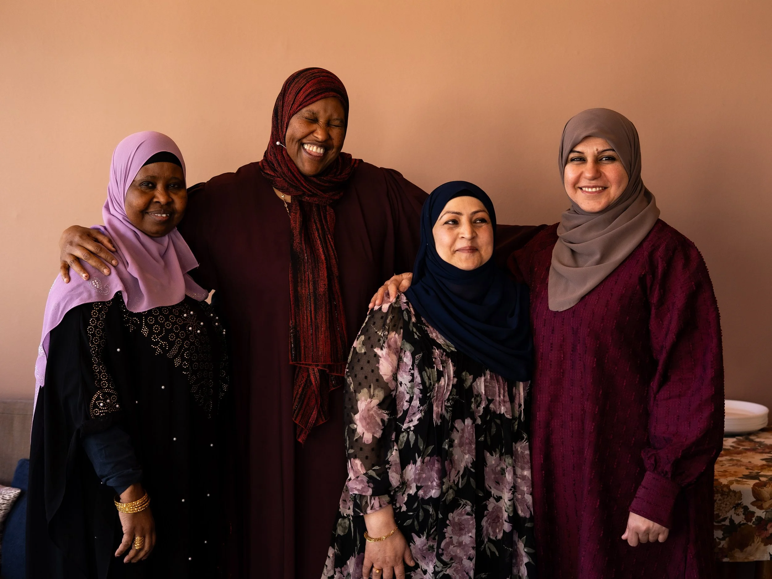 Fyra kvinnor med hijab som ler och står nära varandra, mot en neutral bakgrund.