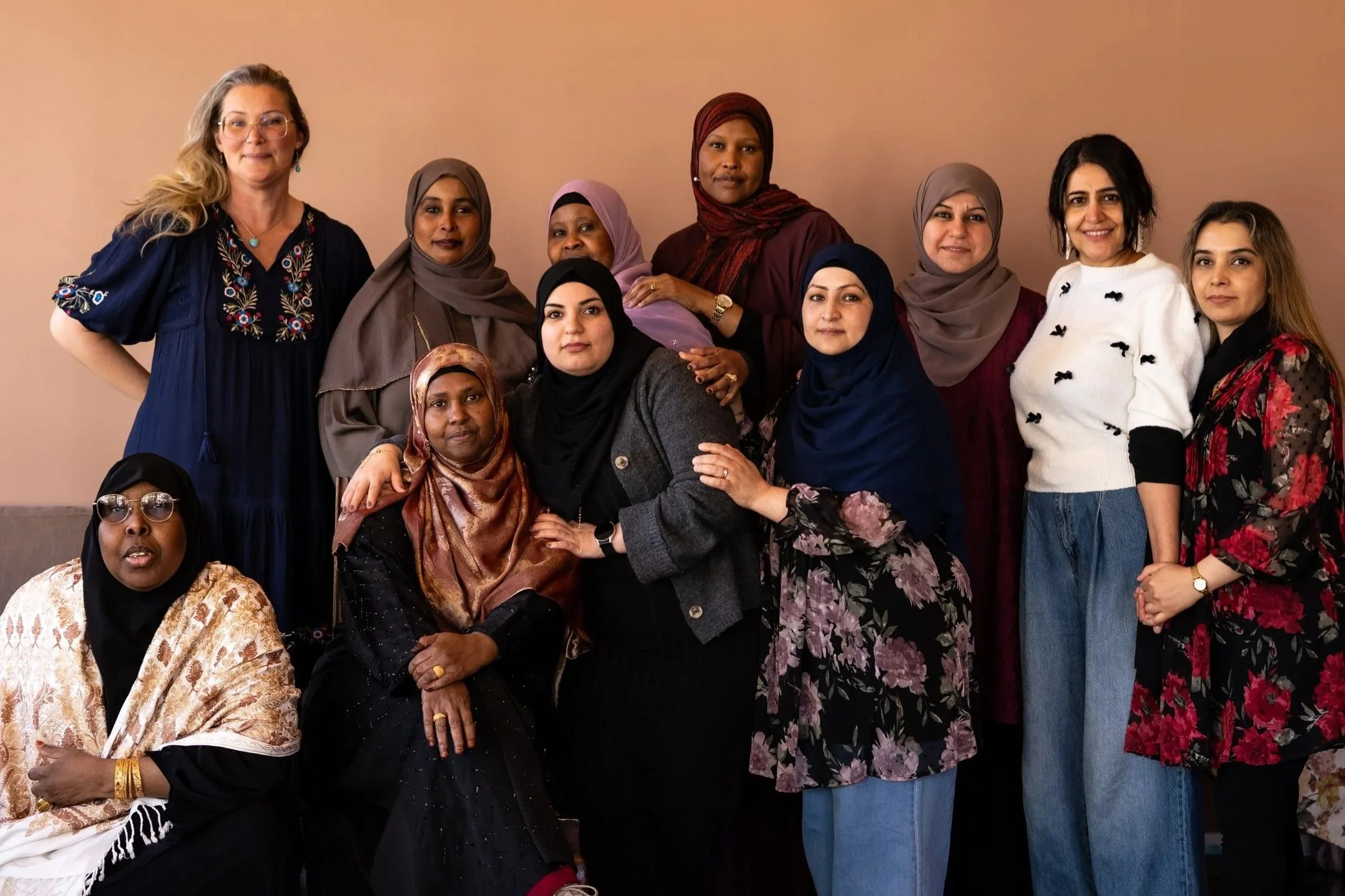 Grupp av kvinnor från olika kulturer och bakgrunder, står tillsammans mot en beige vägg, vissa kvinnor bär hijab, andra inte. Alla ser glada och sammansvetsade ut.
