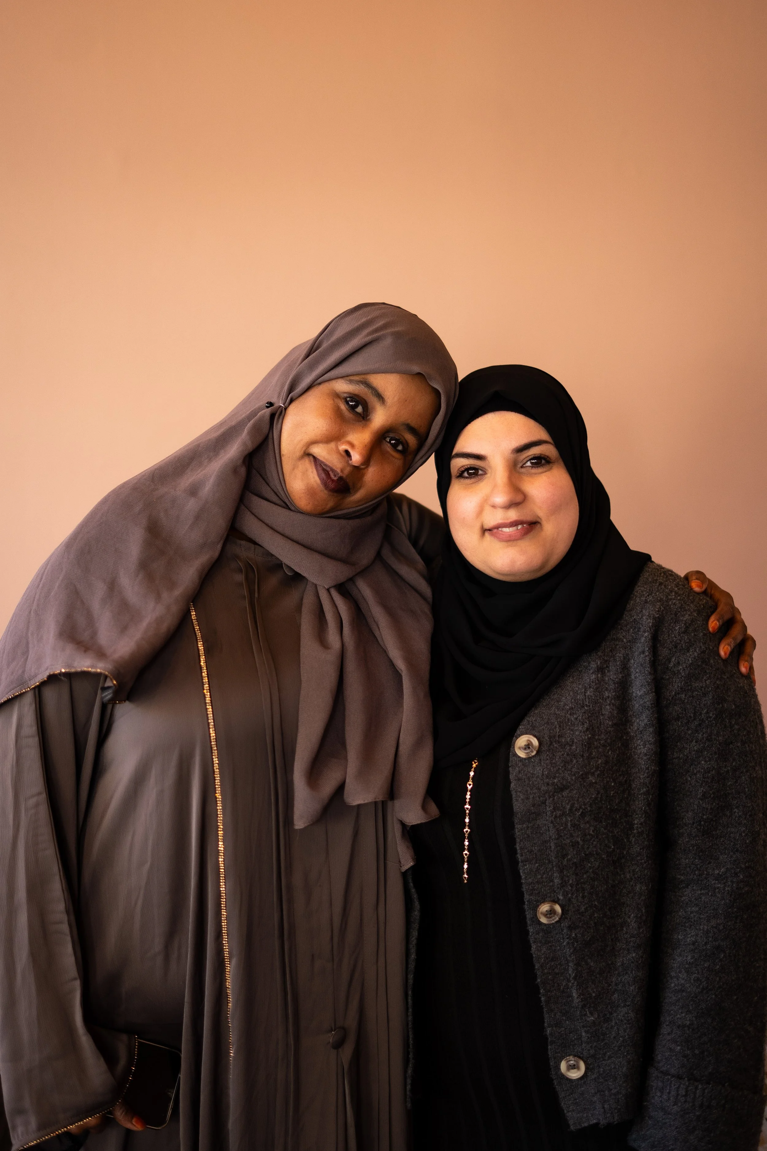 Två kvinnor med hijab, den ena i grå och den andra i svart, står nära varandra och ler mot kameran.