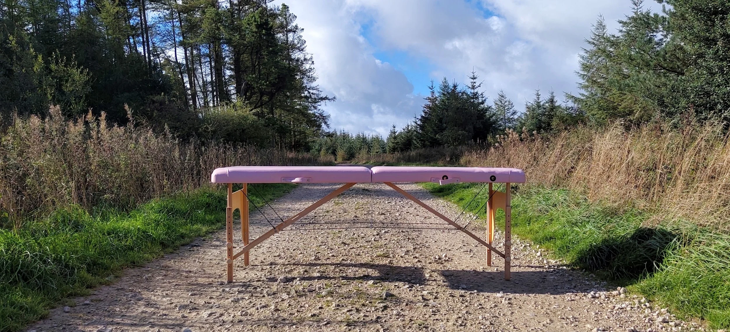 Pink sports massage table set up on a dirt trail in a forested area under a partly cloudy sky.