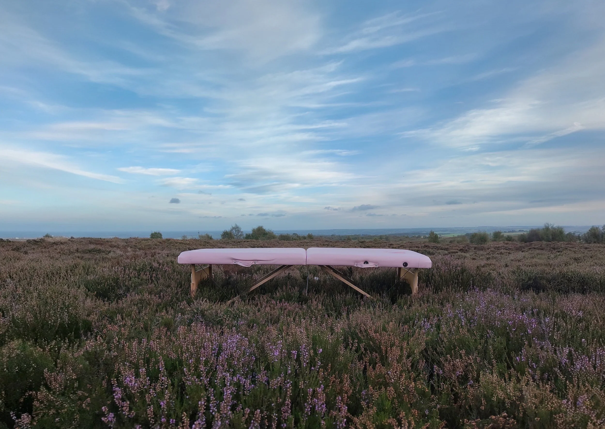 A pink sports massage table set in a field of purple and pink flowers under a partly cloudy sky. Sports Massage available in Otley in evenings and Weekends
