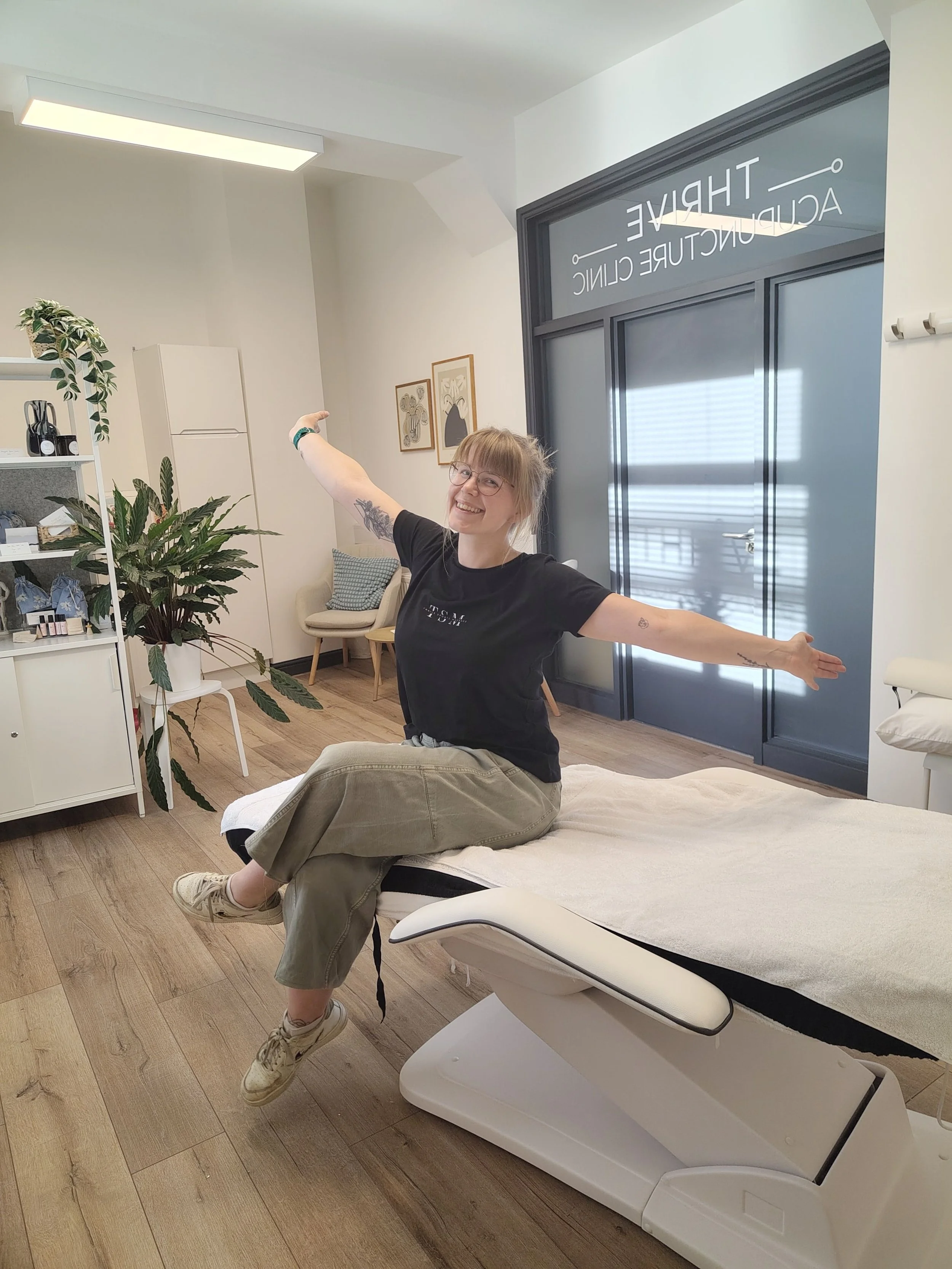 A woman sitting cross-legged on a sports massage bed table in a doctor's office, smiling and stretching with arms wide open. Sports Massage available in Otley in evenings and Weekends