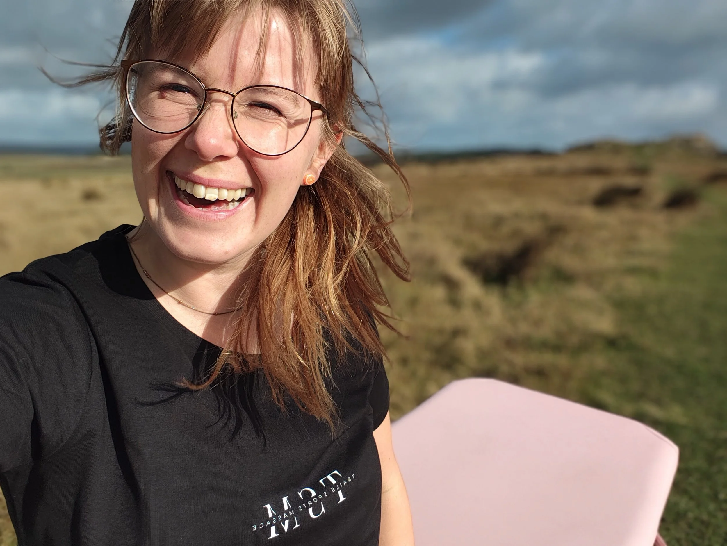 A smiling woman with glasses and earrings, outdoors with cloudy sky and grassy landscape.
