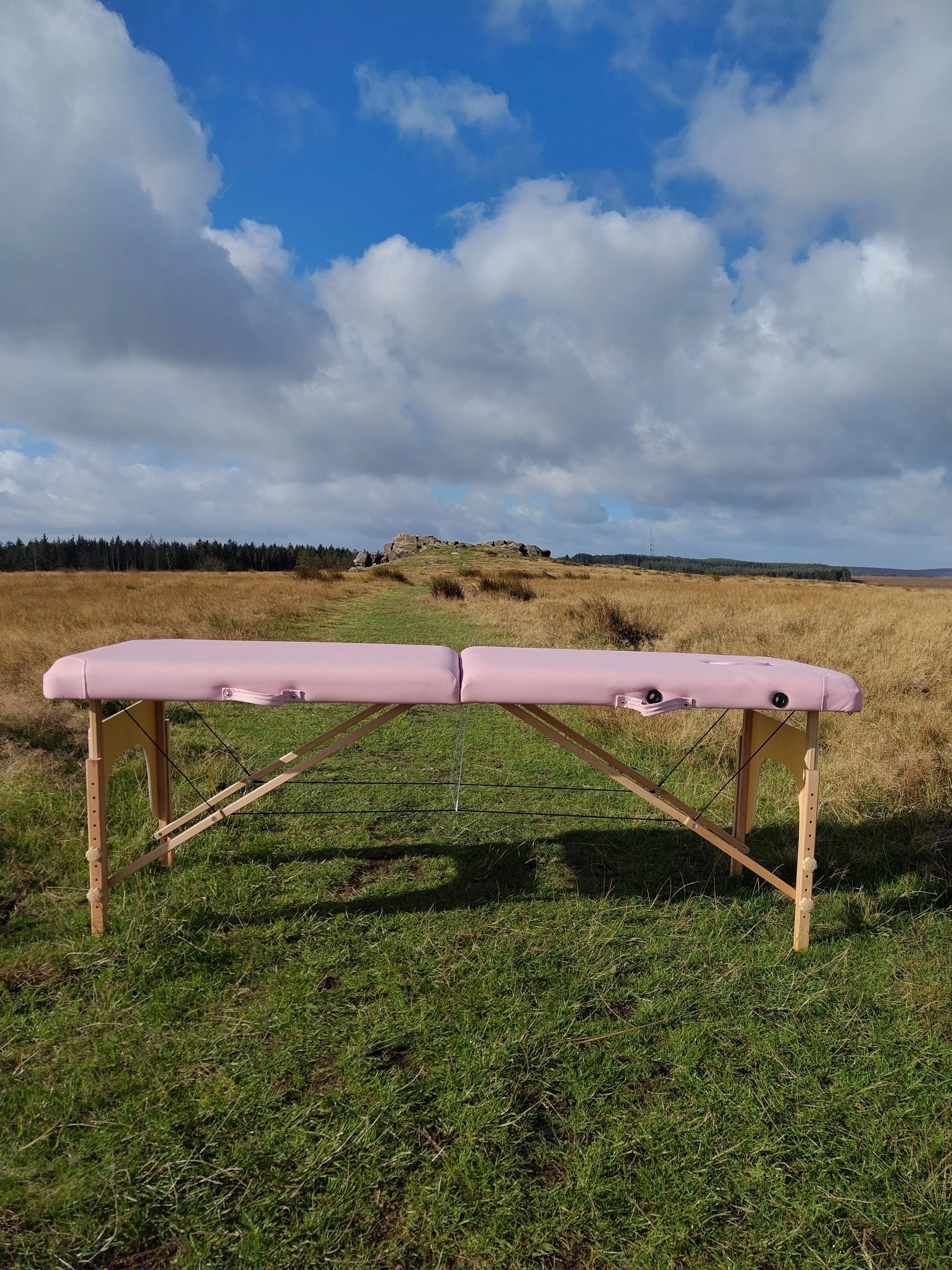 A sports massage table with pink padding set up outdoors in a grassy field, with a landscape of blue sky, clouds, trees, and rocky formation in the background. Sports Massage available in Otley in evenings and Weekends