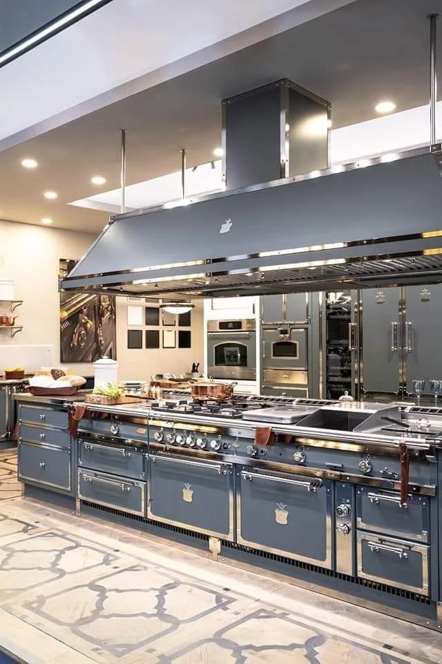 Modern kitchen with multiple ovens, a large stove, metallic and blue cabinetry, and a large industrial range hood.