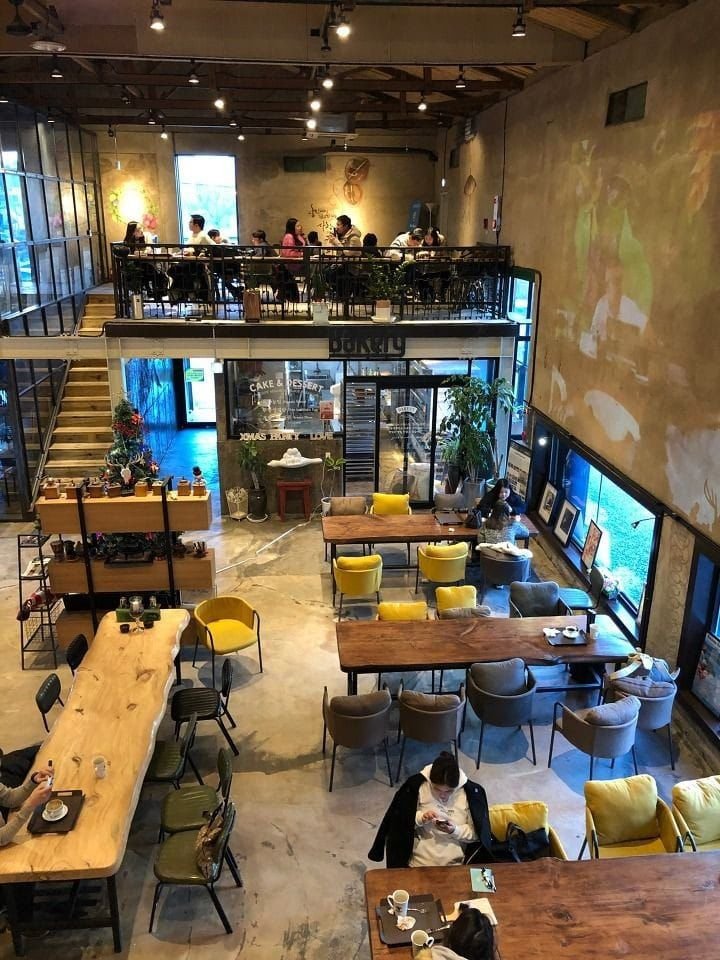 A modern two-story cafe with seating on the ground floor and upper level, decorated with colorful chairs, wooden tables, and potted plants, with people enjoying their time.