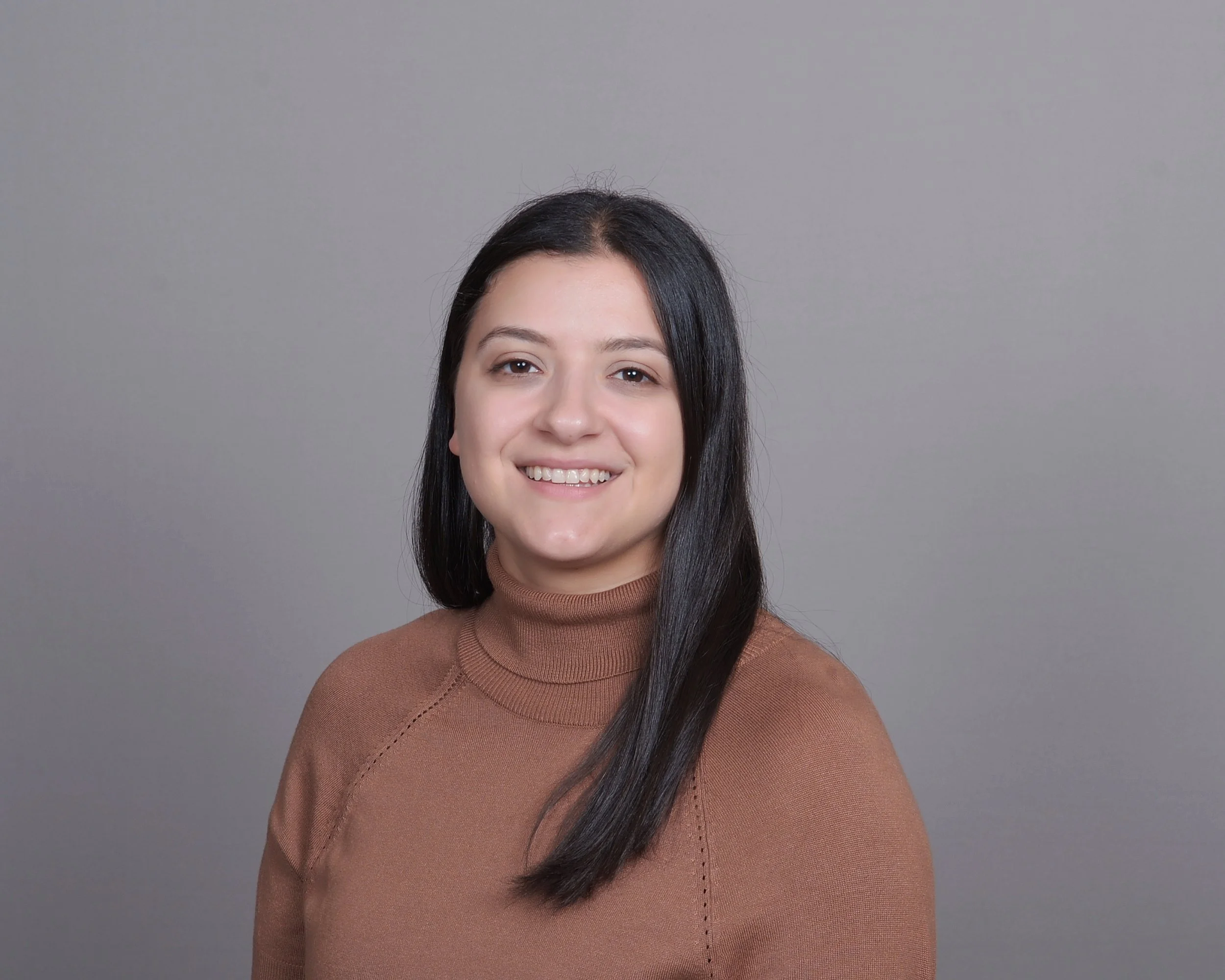A young woman with long dark hair, wearing a brown turtleneck sweater, smiling, standing against a light gray background.