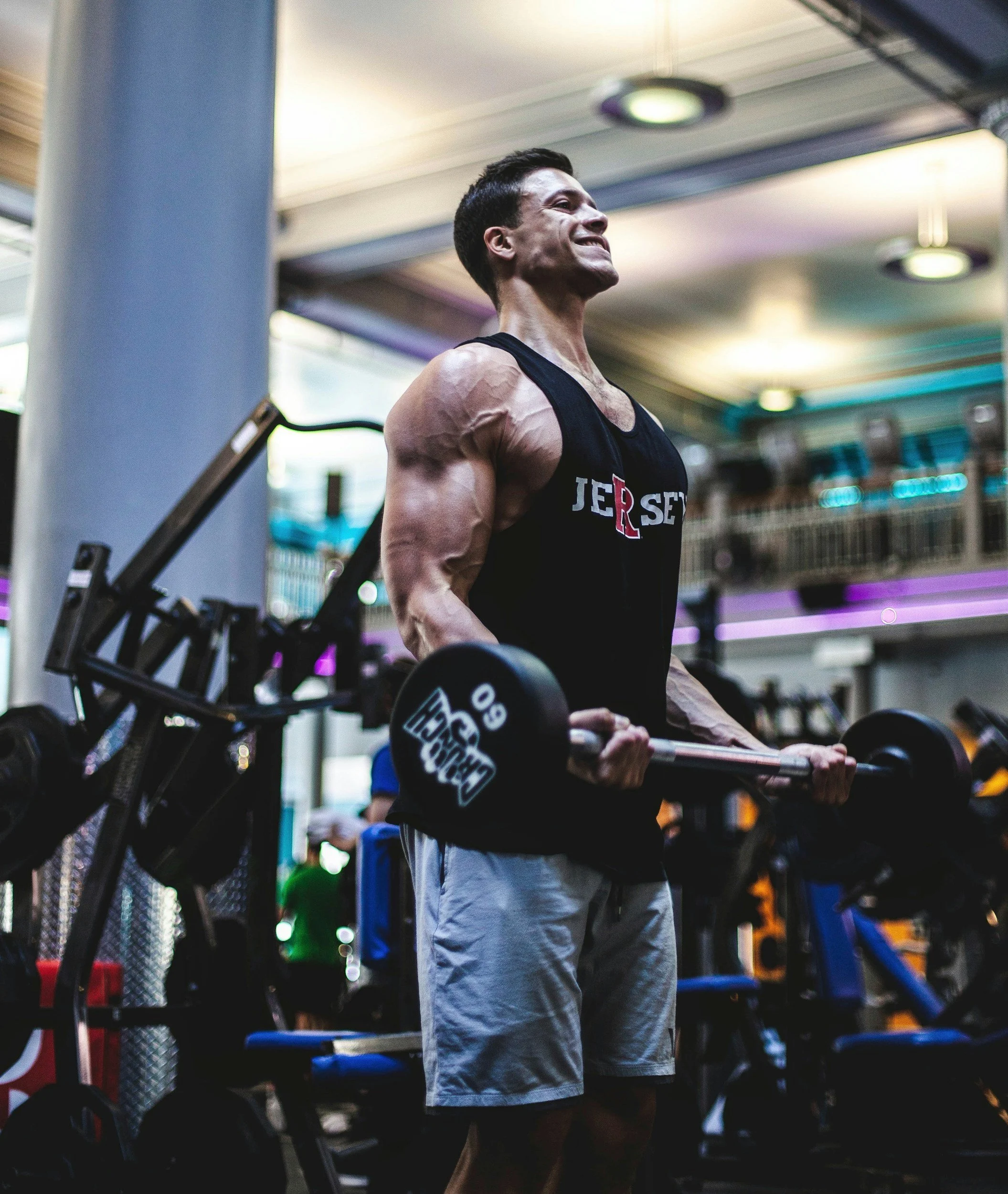 A young man with short dark hair smiling while lifting a barbell in a gym. He is wearing a black sleeveless shirt and light-colored shorts. The gym has weights, exercise equipment, and colorful lighting in the background.