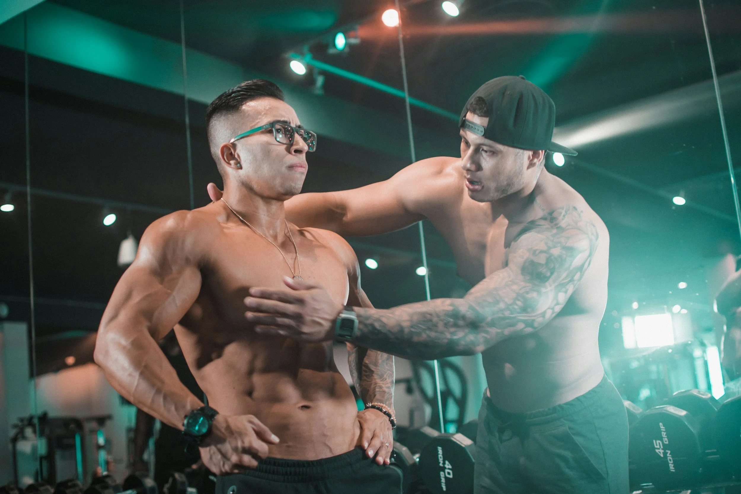 Two shirtless men are in a gym with mirrors and weights. One man, with glasses and a necklace, looks serious. The other man, with tattoos and a cap backwards, is helping him with his chest.