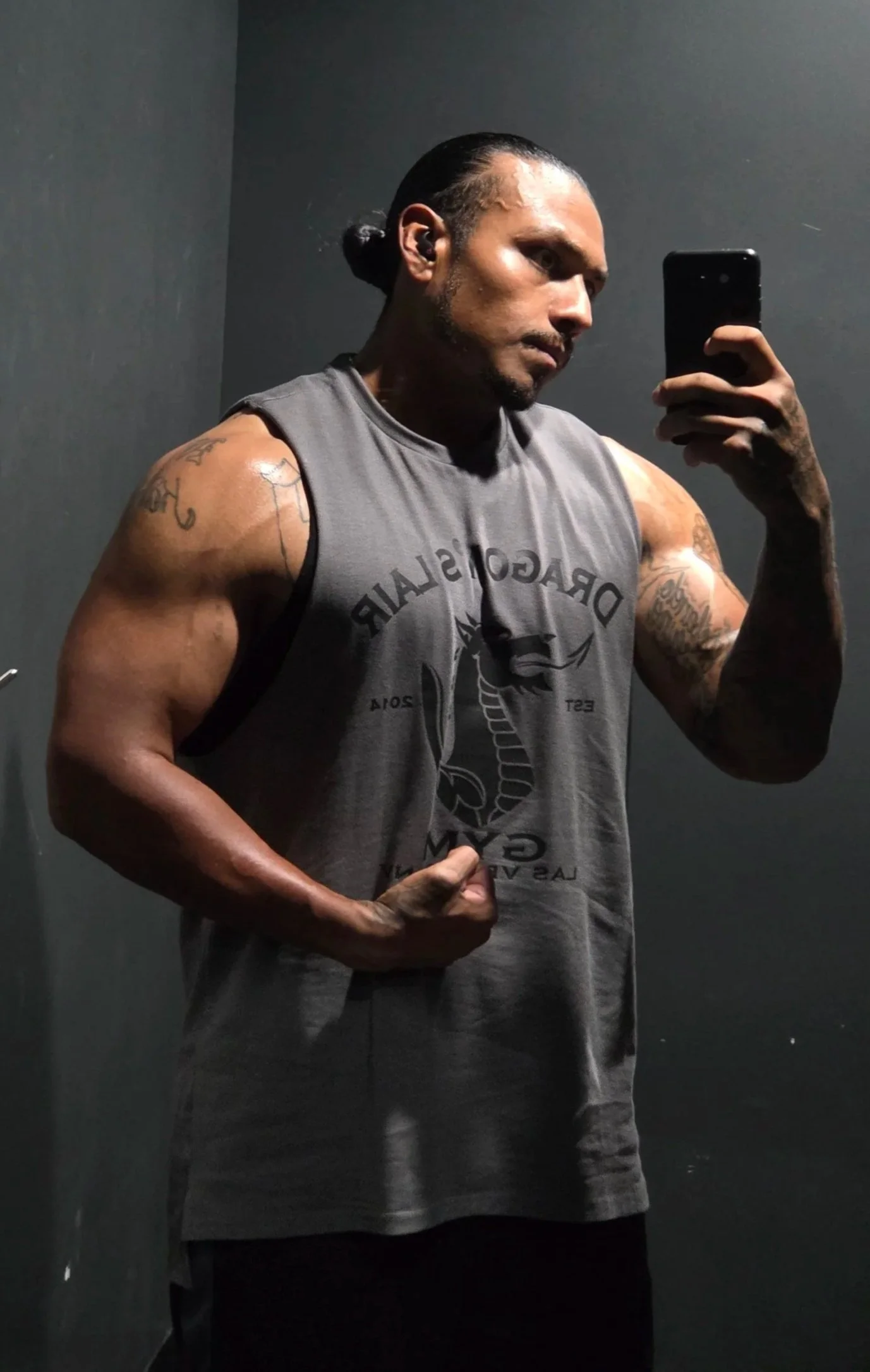 A muscular man taking a mirror selfie in a gym locker room, flexing his right bicep, wearing a sleeveless gray T-shirt and black shorts, with tattoos on his arms and slicked-back hair.