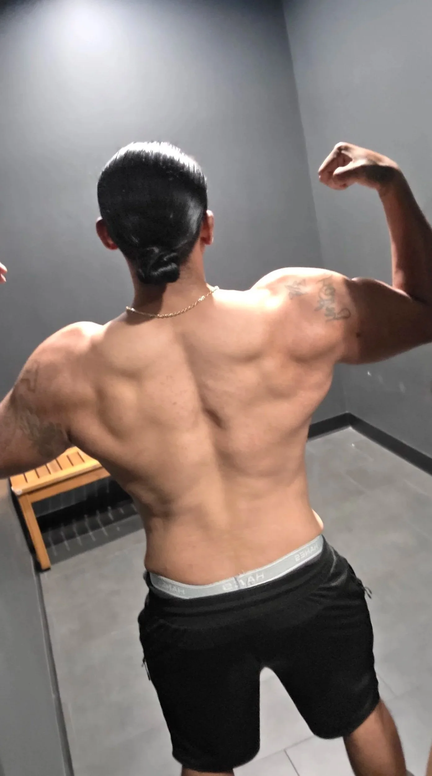 A shirtless man with tattooed arms flexing his muscles in front of a mirror in a room with gray walls and a wooden bench.