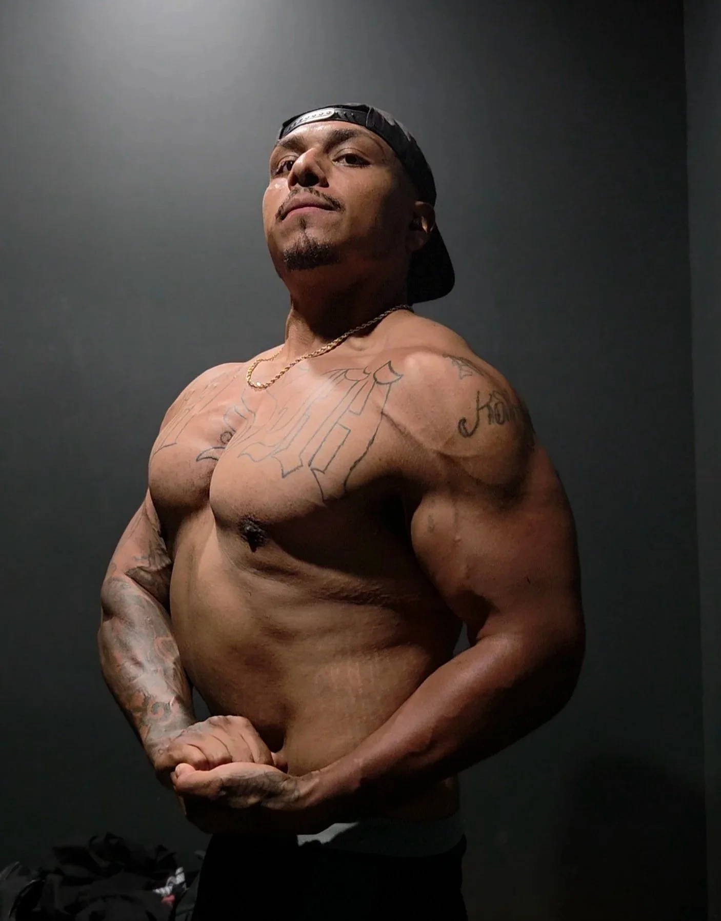 A muscular man with tattoos wearing a backward cap and necklace, posing shirtless against a dark background.