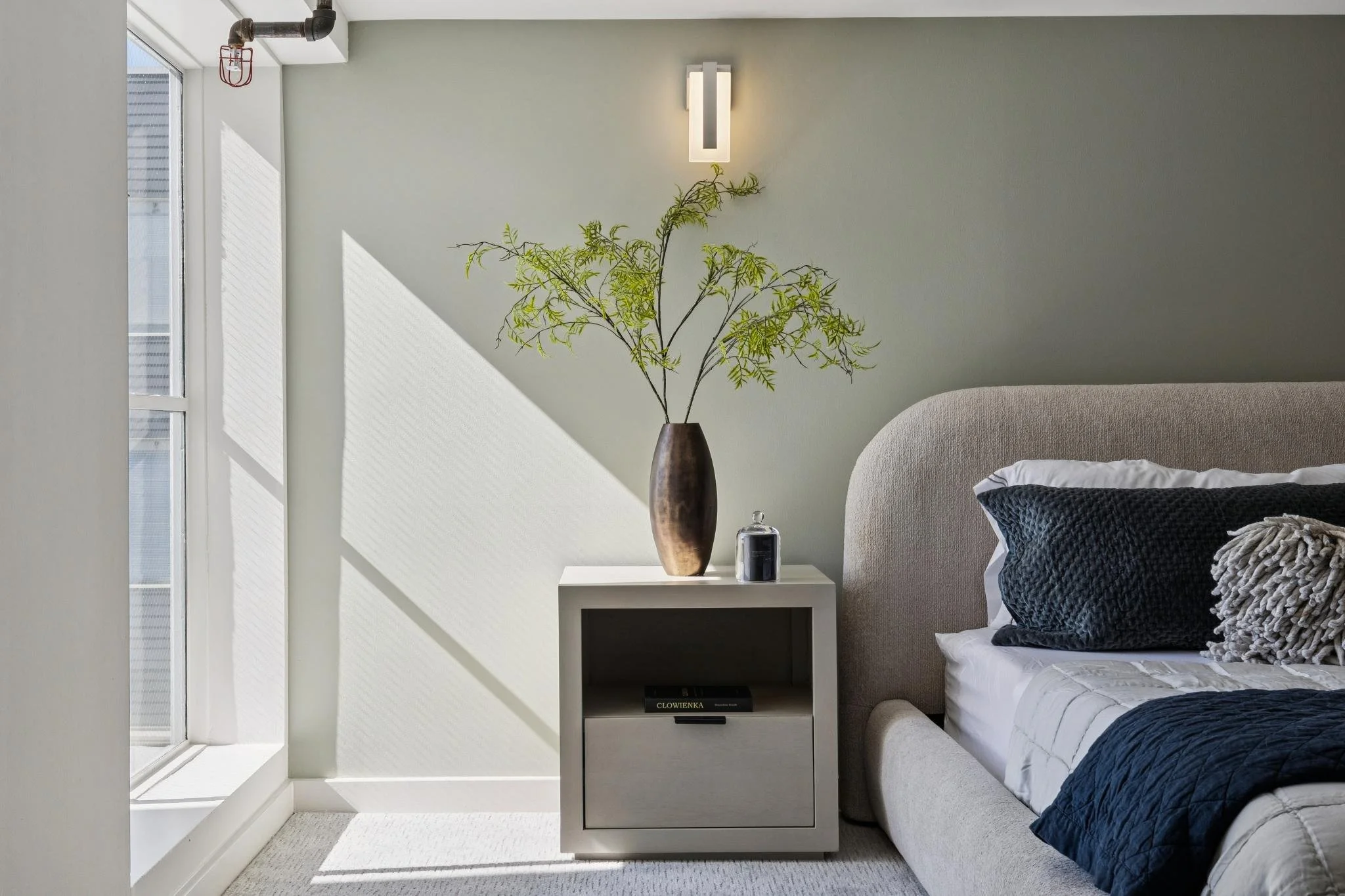 A bedroom corner with a window, a beige bed with navy blue and gray pillows, a nightstand with a tall vase with green branches and a small jar, and a wall-mounted light. Natural sunlight streams through the window creating shadows on the wall.