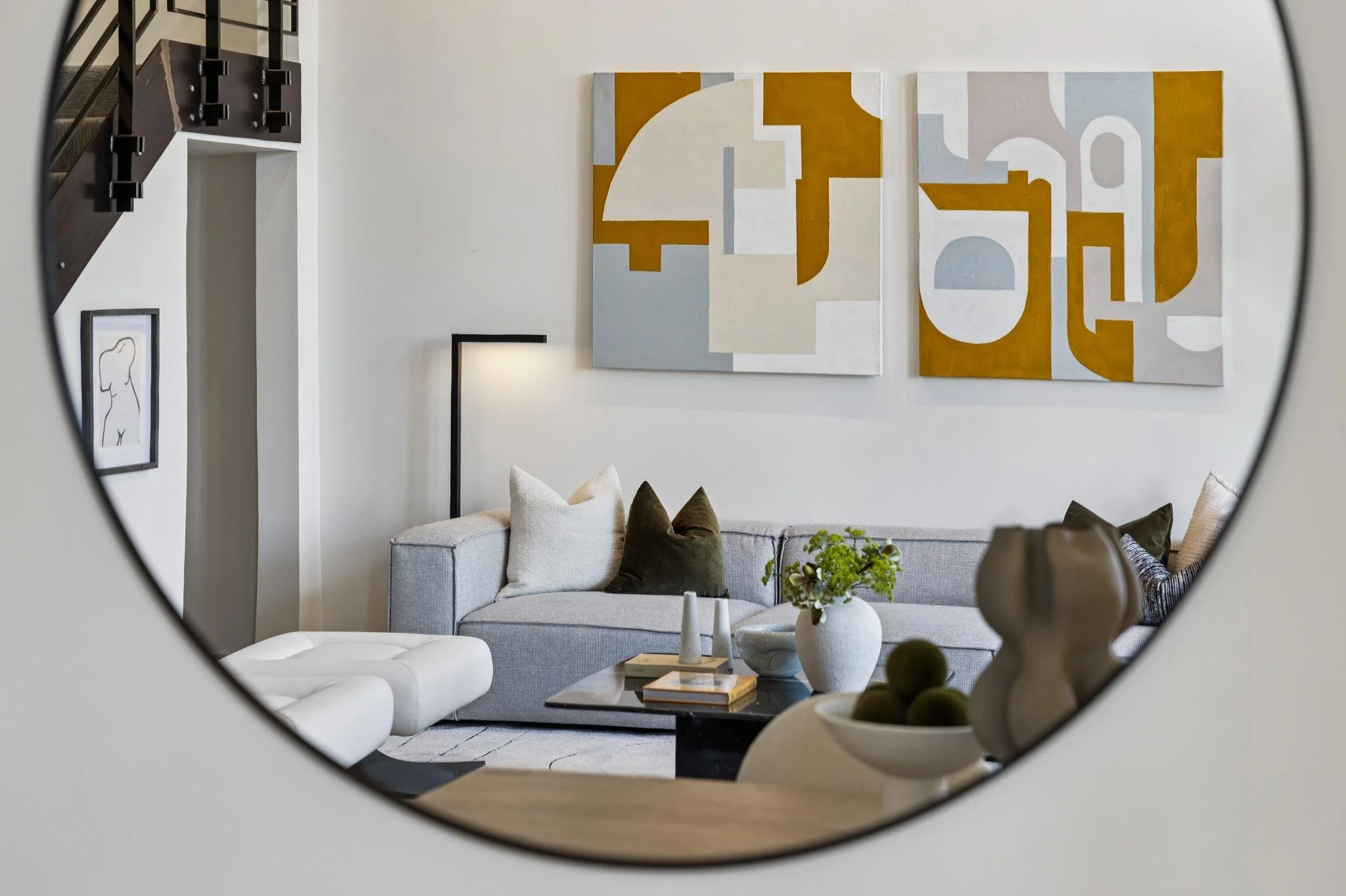 Living room with a light grey sofa, black coffee table, and modern abstract paintings on white walls, as seen through a circular mirror.