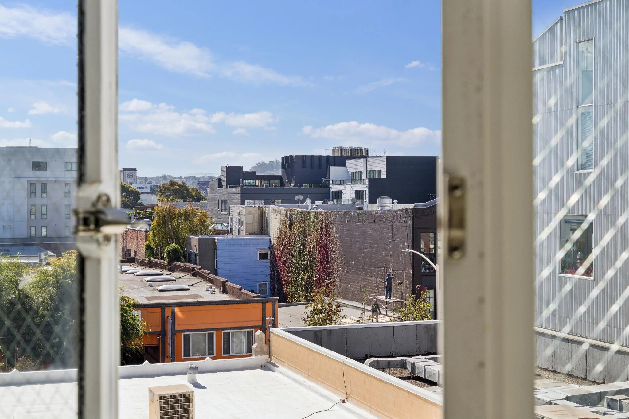 View of a cityscape through a window with white frames, overlooking rooftops, trees, and modern buildings with blue sky and clouds.