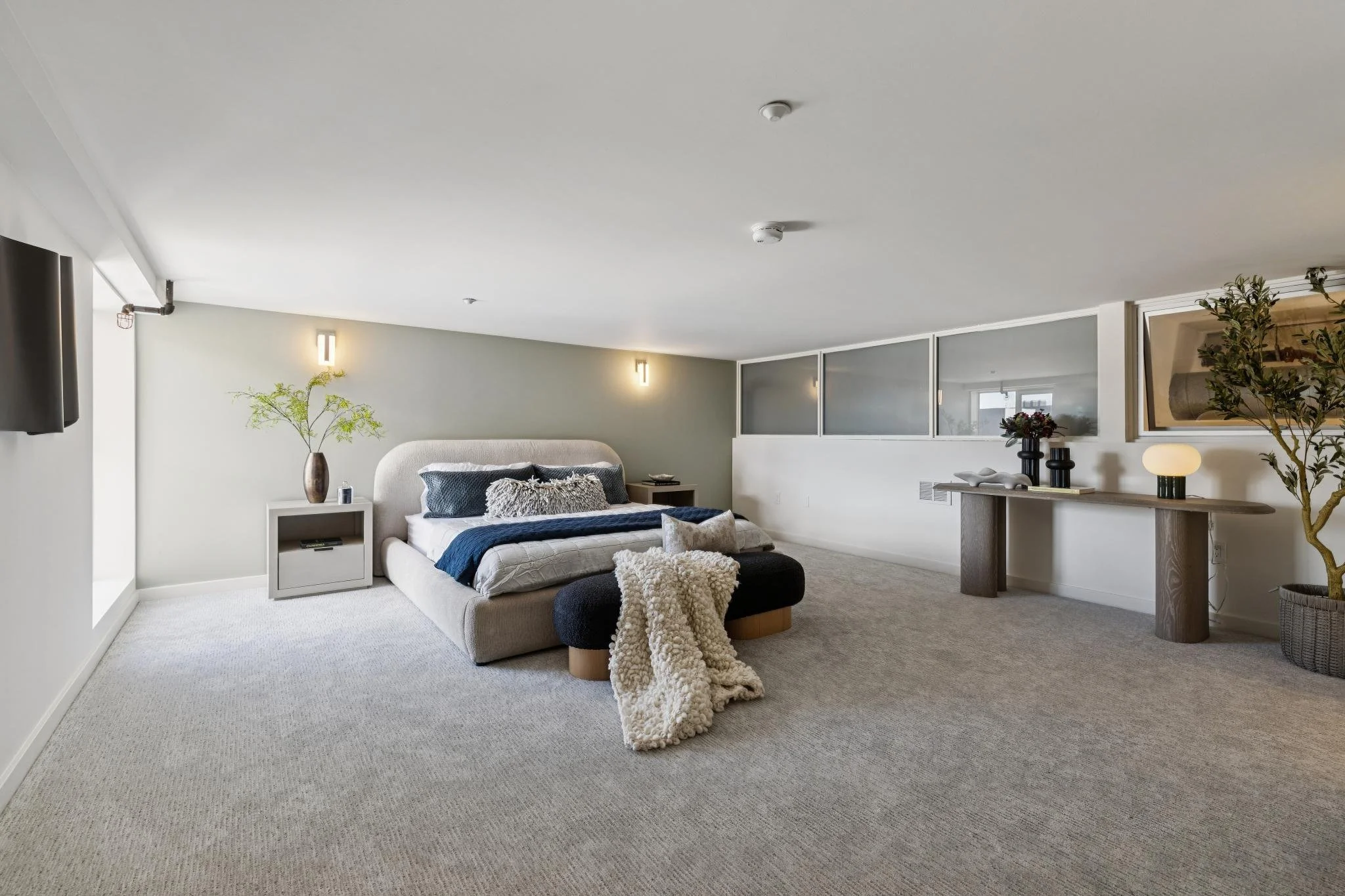 Modern bedroom with beige upholstered bed, blue bedding, cream throw blanket, side table with plant and small decor, wall-mounted TV, and decorative plants, in a room with beige carpet and soft lighting.