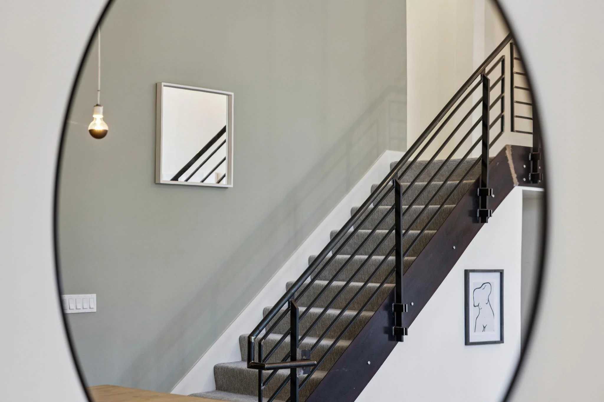 A staircase with dark wooden steps and black metal railing, framed by a circular mirror, with framed artwork on a gray wall, a hanging light bulb, and a white electrical outlet visible.