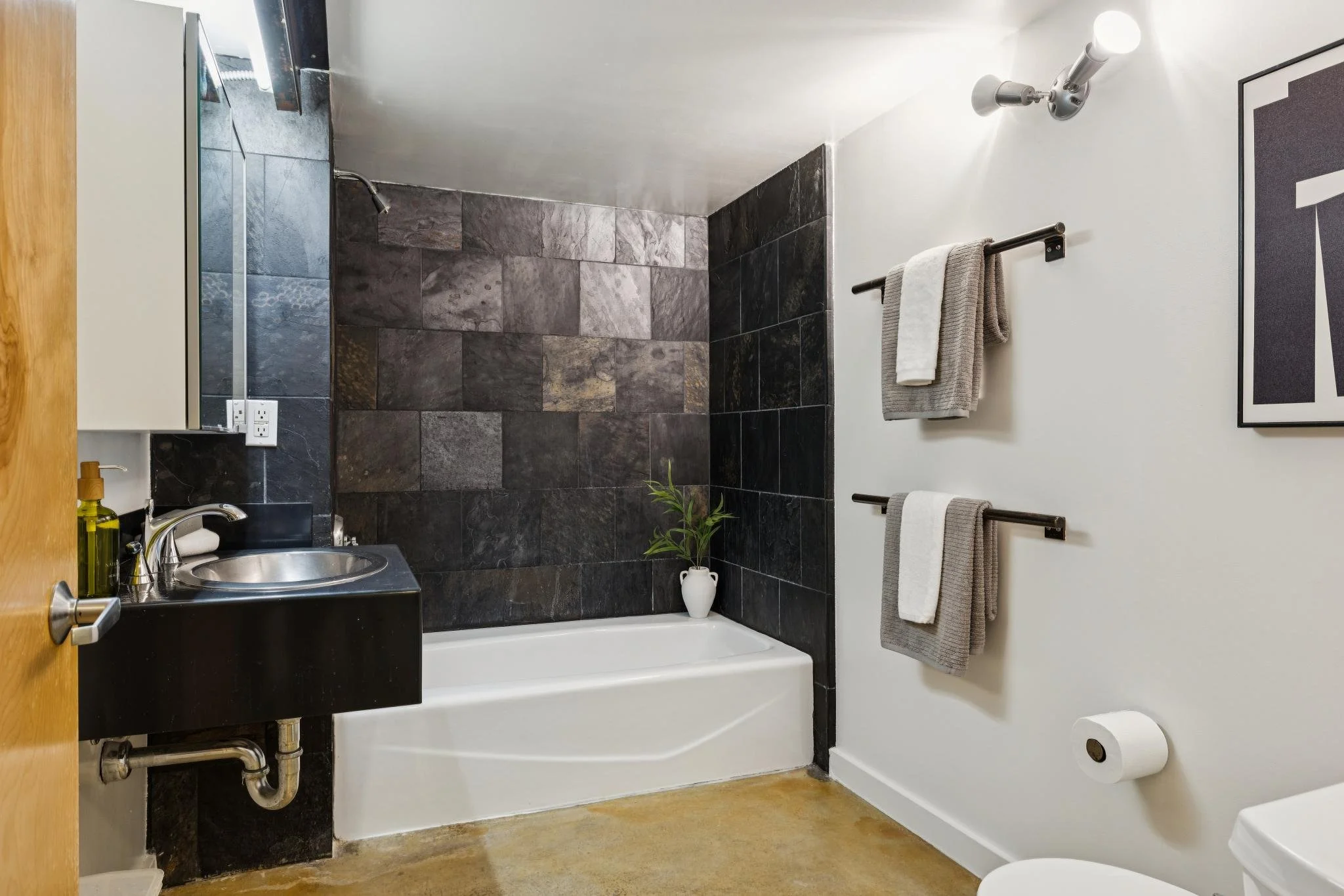 A bathroom with a black vanity and stainless steel sink, black tiled shower area, white walls, towel rack with two gray towels, a framed piece of art, and a plant in a white vase.