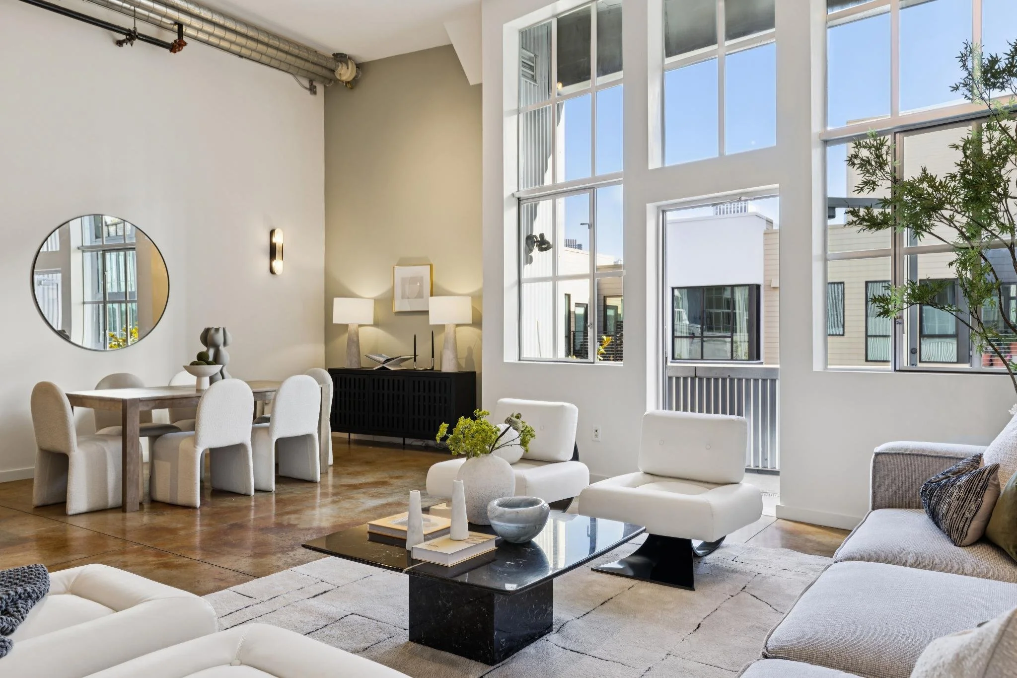 Modern living room with large windows, beige chairs, a black coffee table with decorative objects, and a white sofa with throw pillows. There is a dining area with a round table and upholstered chairs, a wall mirror, and minimalist lamps and artwork 