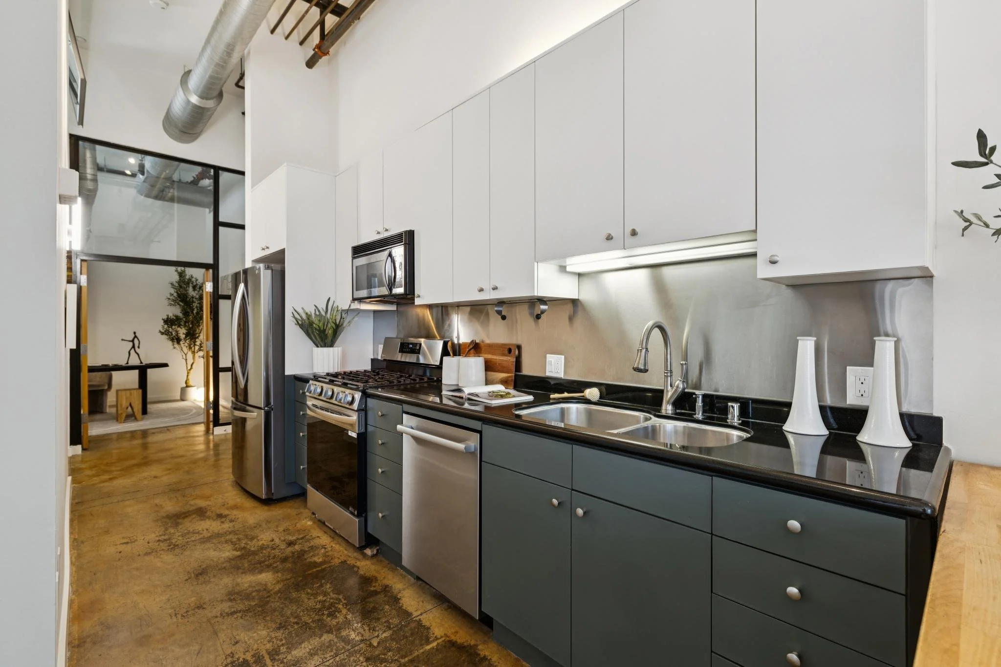Modern kitchen with white upper cabinets, dark lower cabinets, stainless steel appliances including a microwave, refrigerator, and oven, a double sink, and decorative white vases on the countertop. Exposed ductwork and a large window are visible, wit