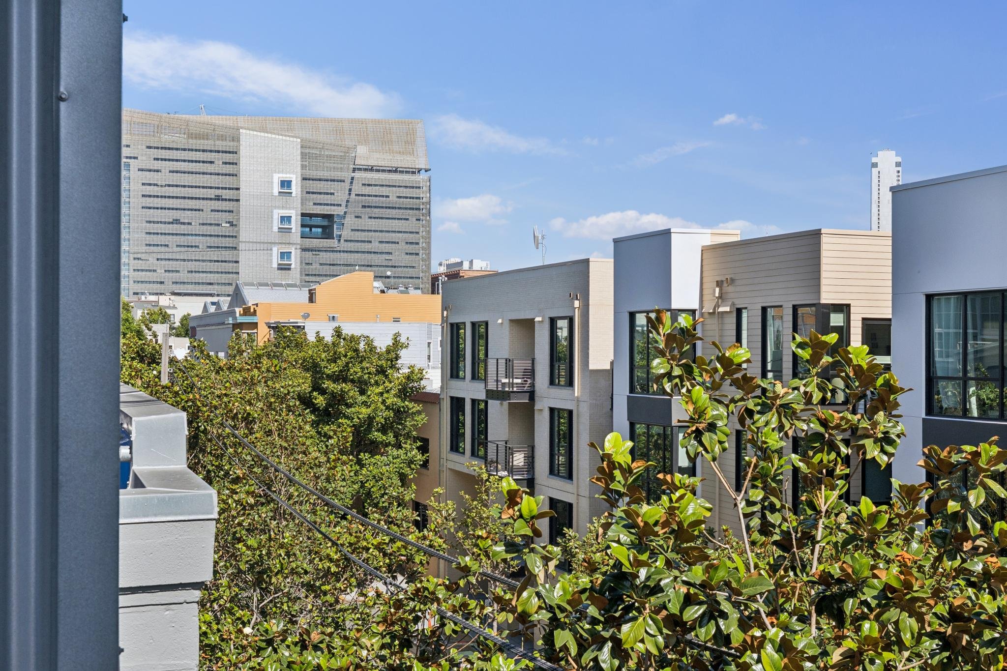 Cityscape featuring modern apartment buildings with trees and a large office building in the background under a blue sky with scattered clouds.