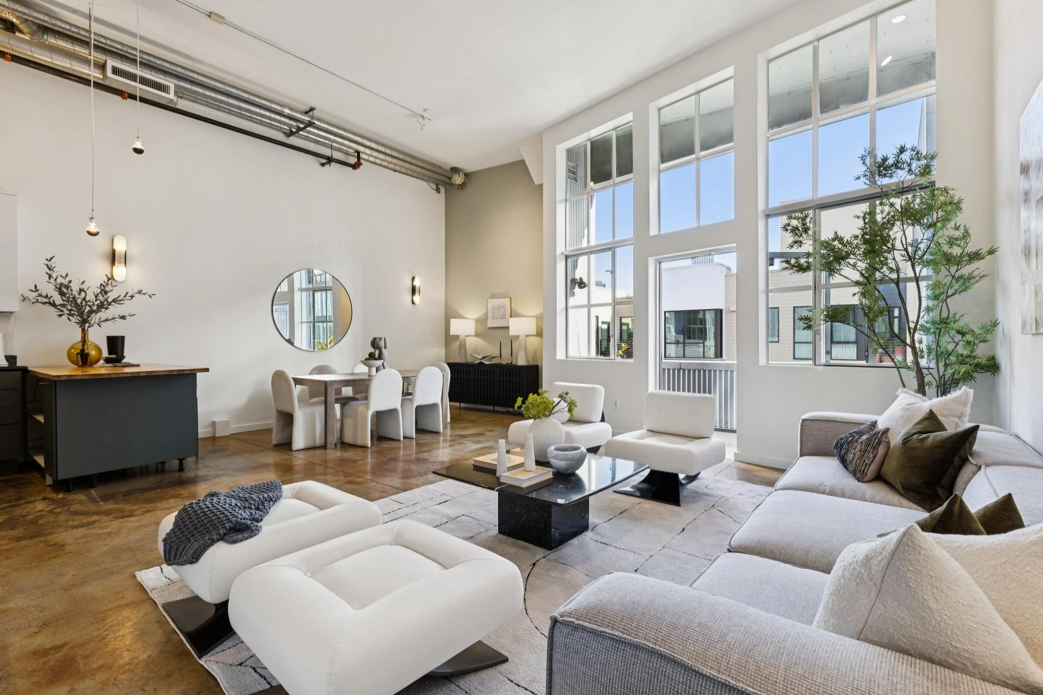 A modern, spacious living room with large windows, white and gray furniture, plants, and decor in neutral tones.