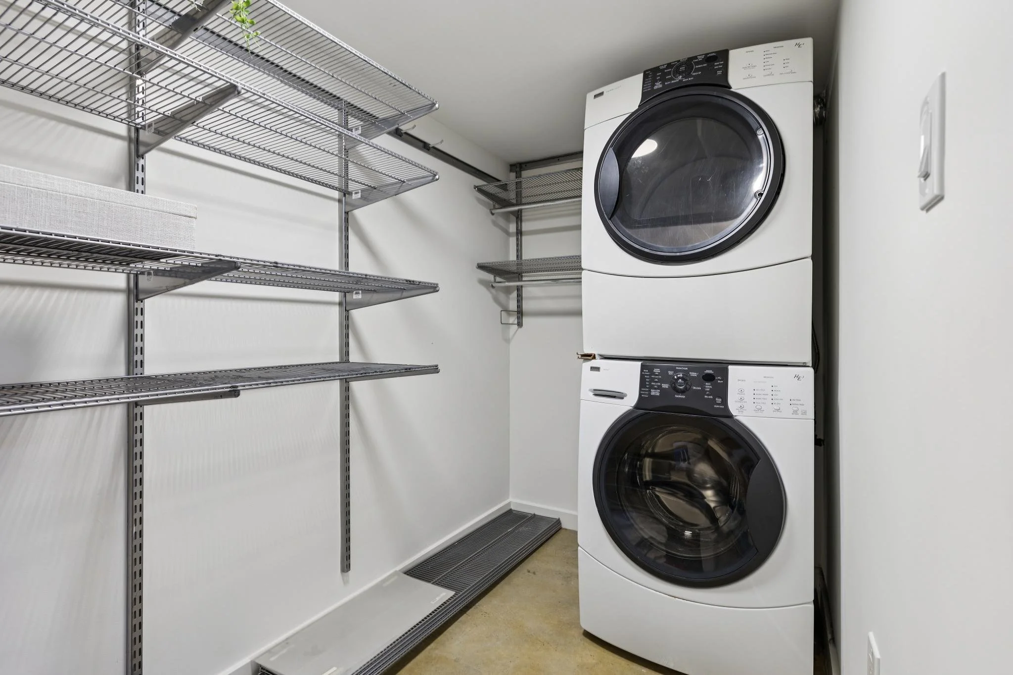Empty laundry closet with wire shelving units, stacked washer and dryer, and a power outlet.