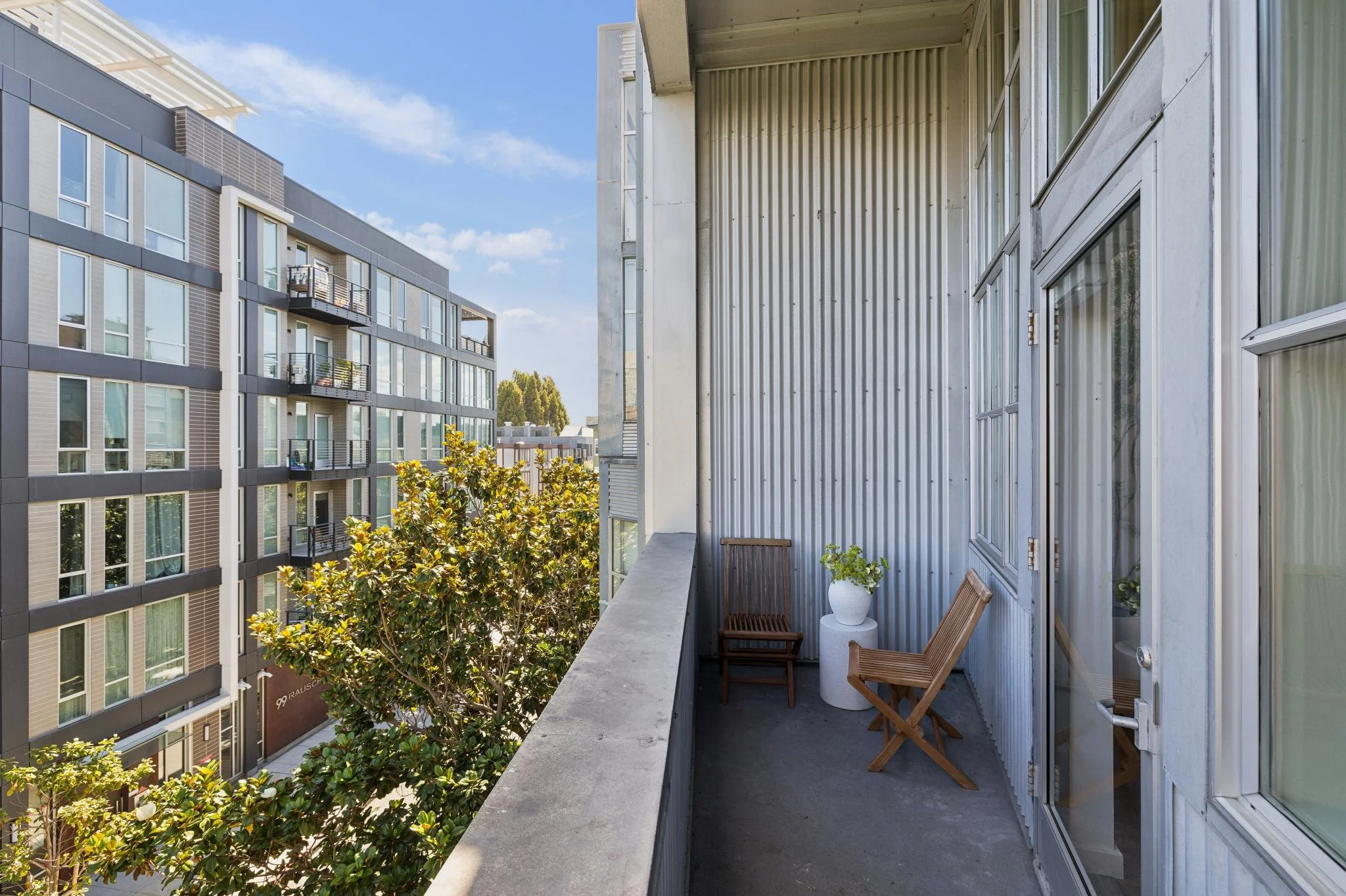 Balcony with wooden chairs and white potted plant, overlooking neighboring modern apartment building with glass windows and balconies, and trees with green foliage.