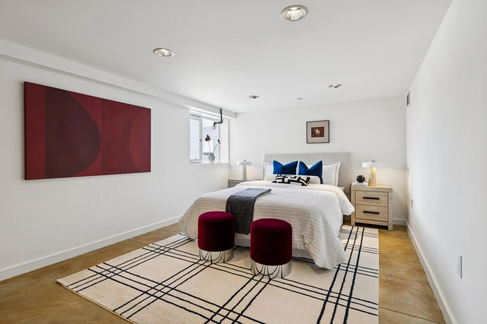 A modern bedroom with white walls, a bed with white bedding, blue pillows, and a patterned black and white rug. There are two red velvet stools in front of the bed, a nightstand with lamps and decor, artwork on the walls, and a window with a vase and