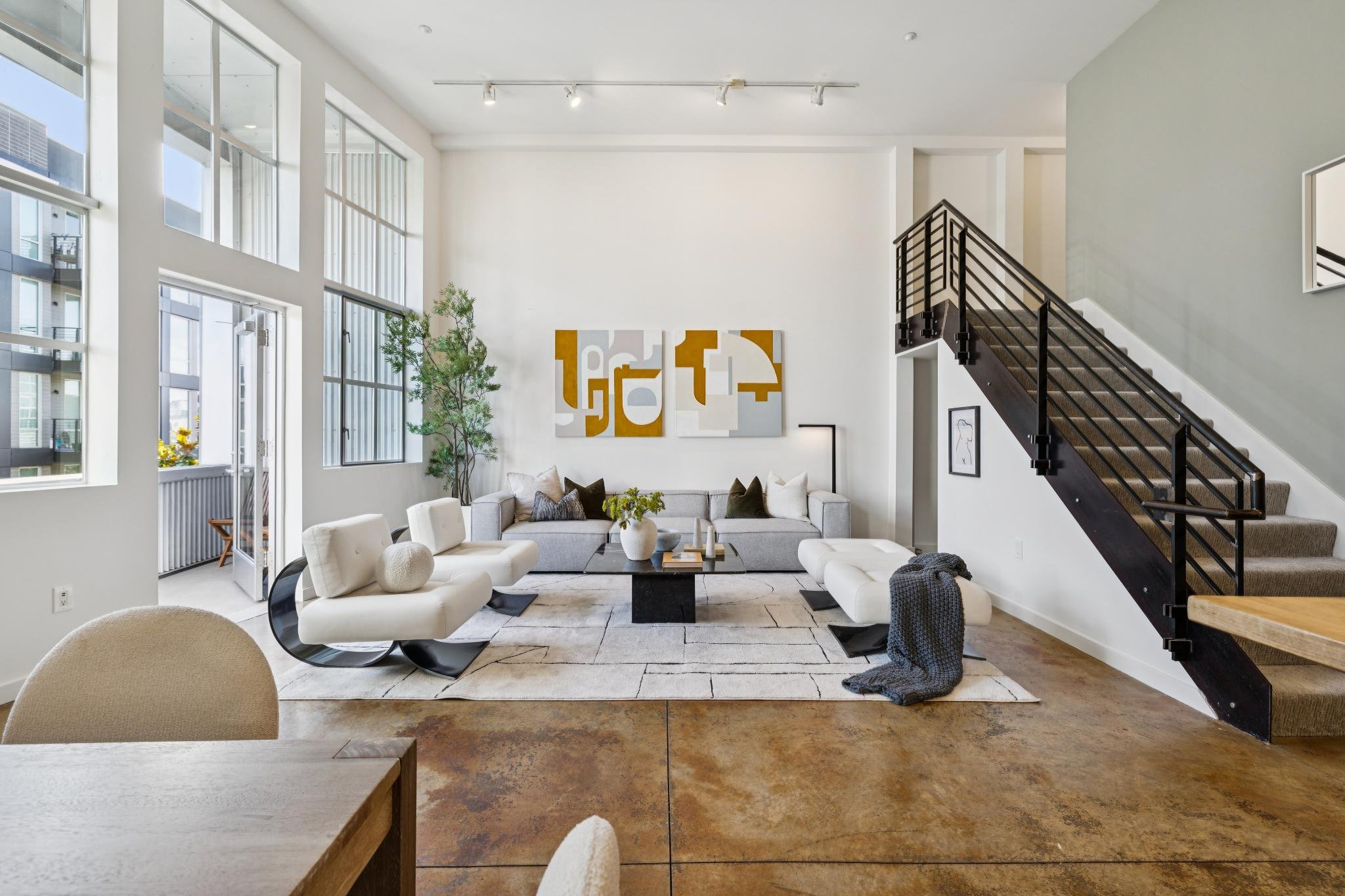 Modern living room with high ceilings, large windows, white walls, abstract artwork, gray sectional sofa, white modern chairs, wooden staircase with black railing, and indoor plants.