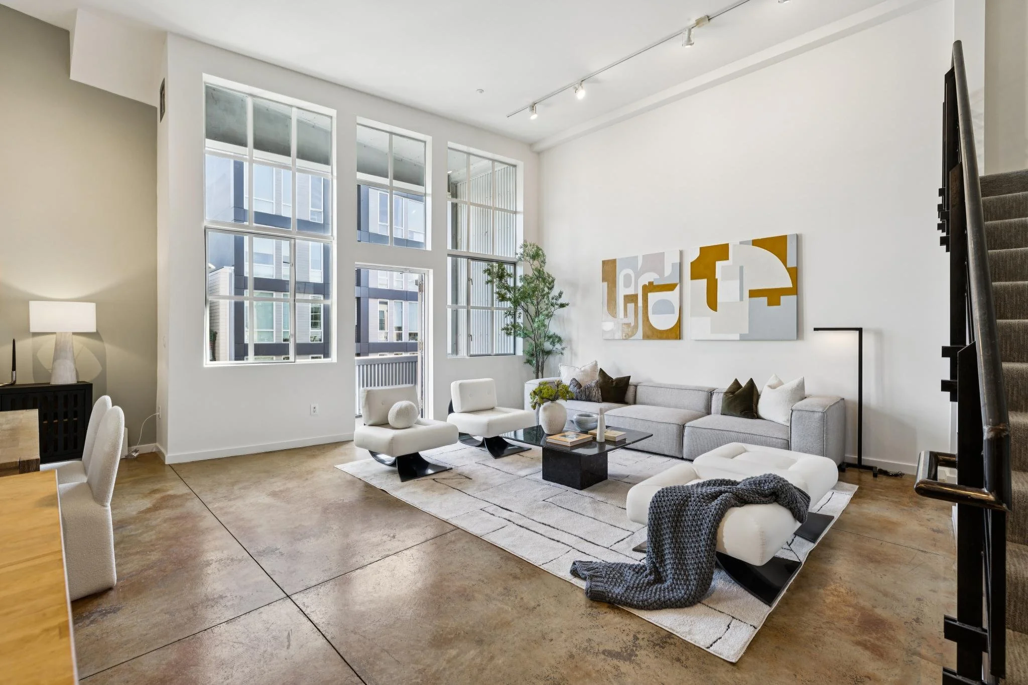 Modern living room with large windows, white walls, abstract wall art, gray sectional sofa, white chairs, black coffee table, green plant, and staircase.
