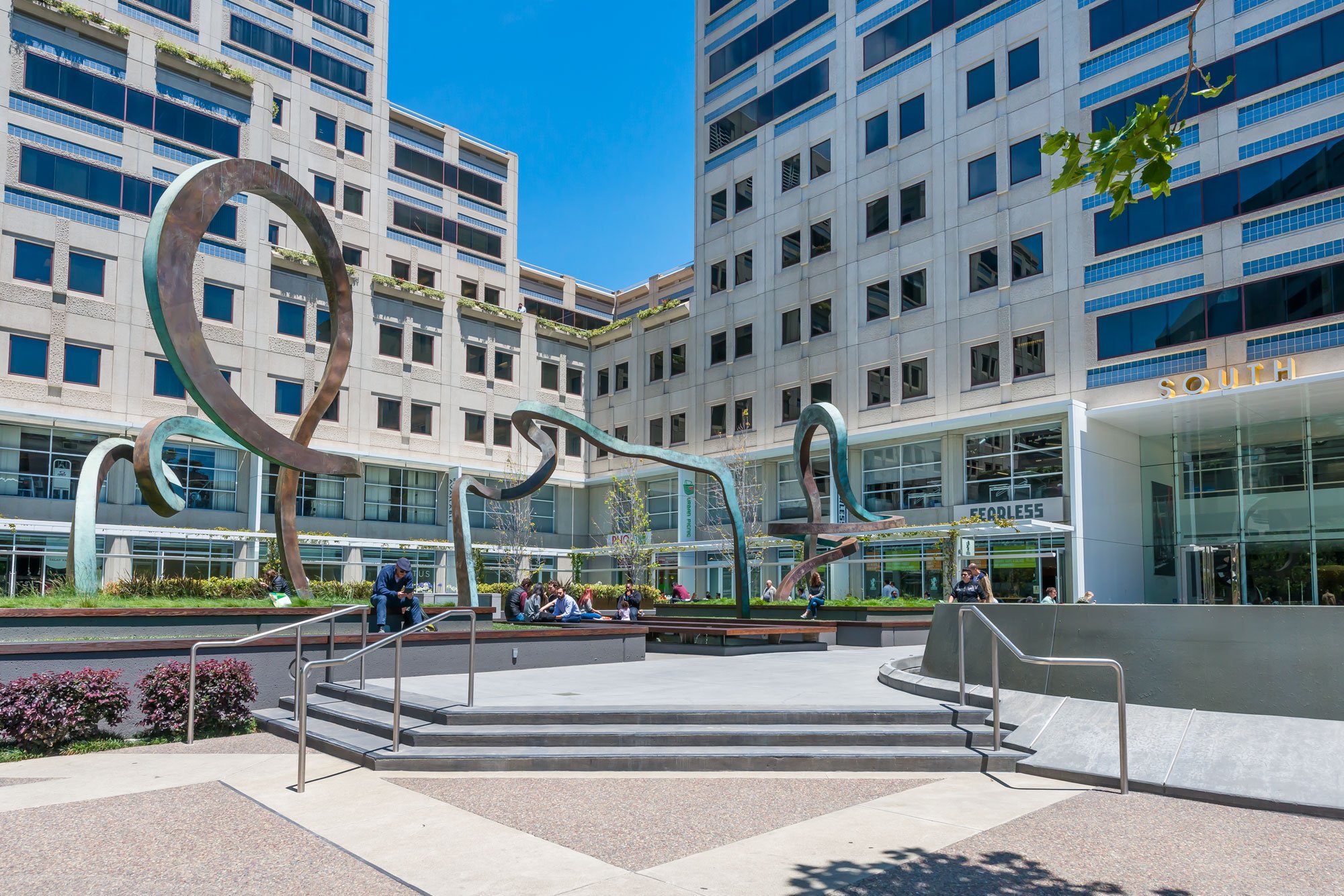A modern city plaza with a multi-story building in the background. The plaza features abstract metal sculptures, benches, and some people sitting and walking. The building has large windows and balconies, with a sign that reads 'SOUTH' and storefront