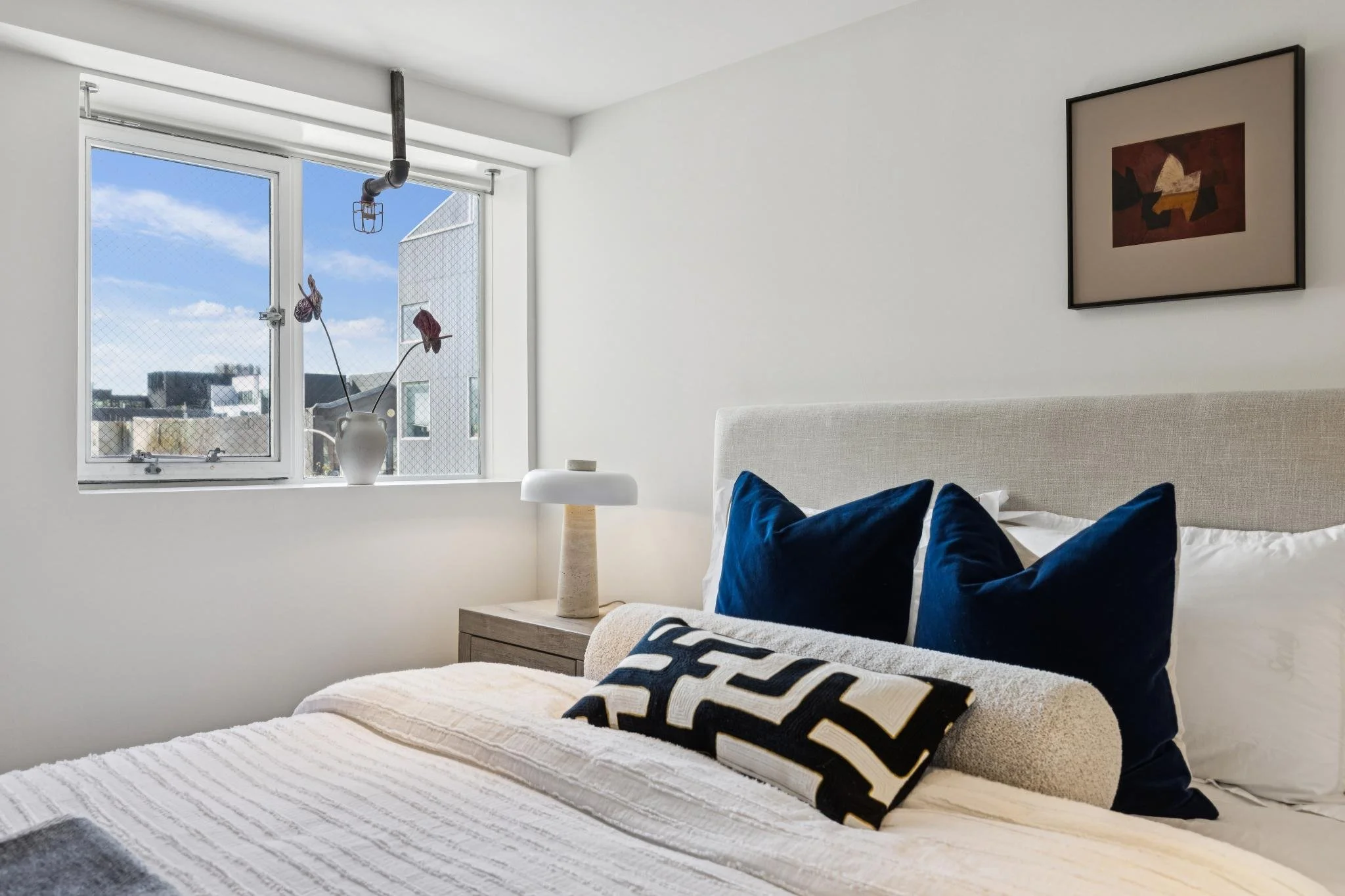 Bedroom with white walls, window with city view, decorative vase on bedside table, blue pillows on bed, framed artwork on wall.