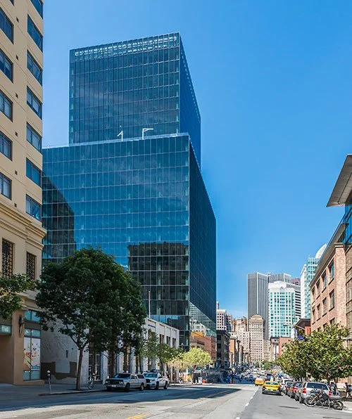 Downtown city street with tall modern glass skyscraper, older buildings, and trees lining the sidewalk.