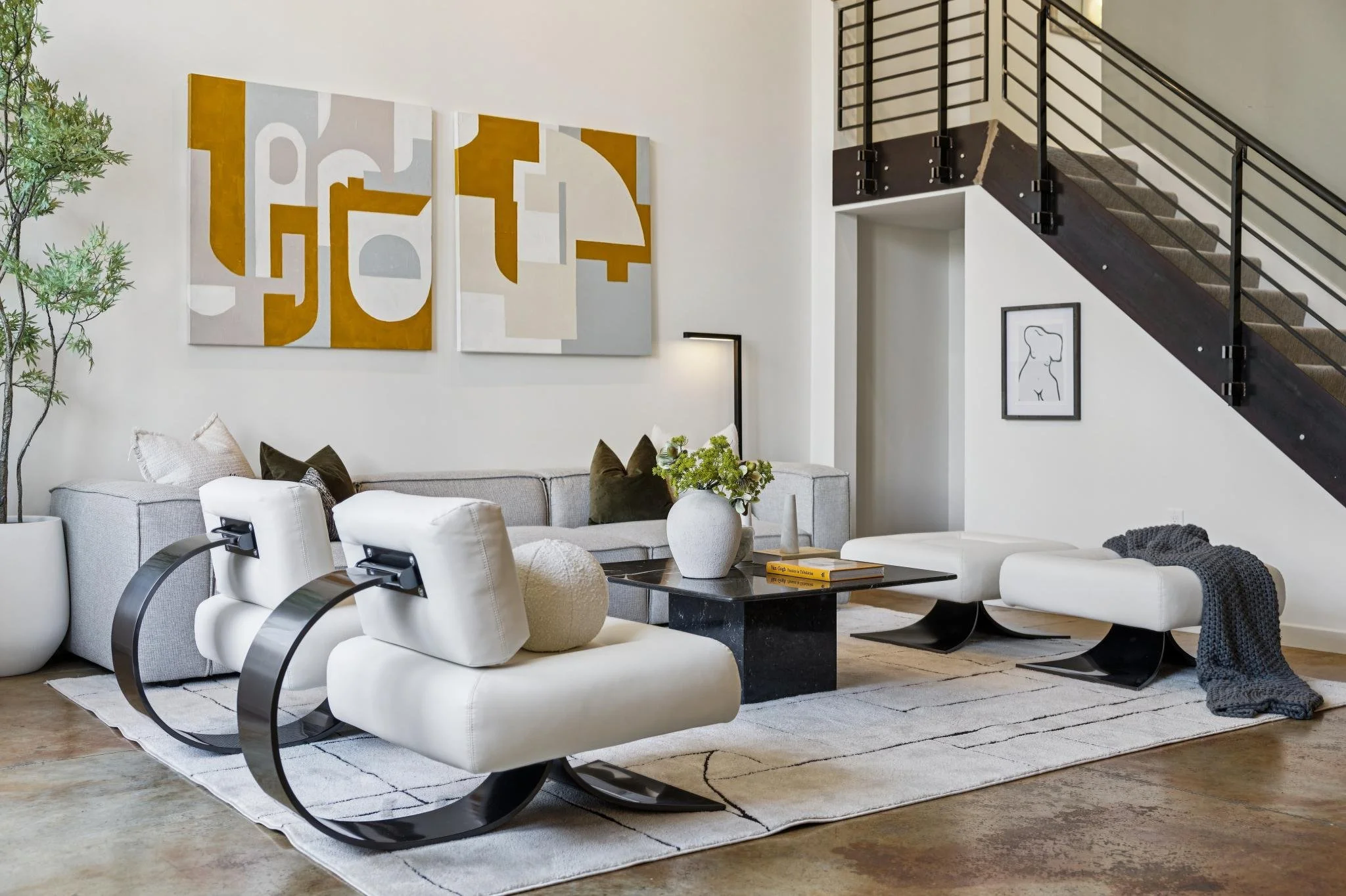 Modern living room with white sectional sofa, black rectangular coffee table, beige armchair with black accents, abstract gold and white wall art, and a staircase with black railing.