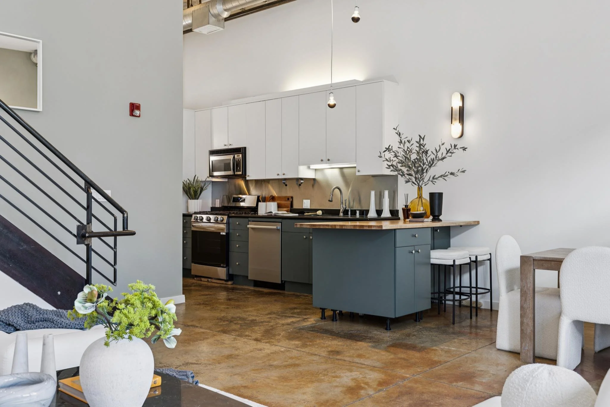 A modern open-concept kitchen and dining area with a white upper cabinet, dark lower cabinet, a stainless steel stove, microwave, and a wooden kitchen island. Decorated with vases, plants, and a wall-mounted light fixture. Part of a living space with