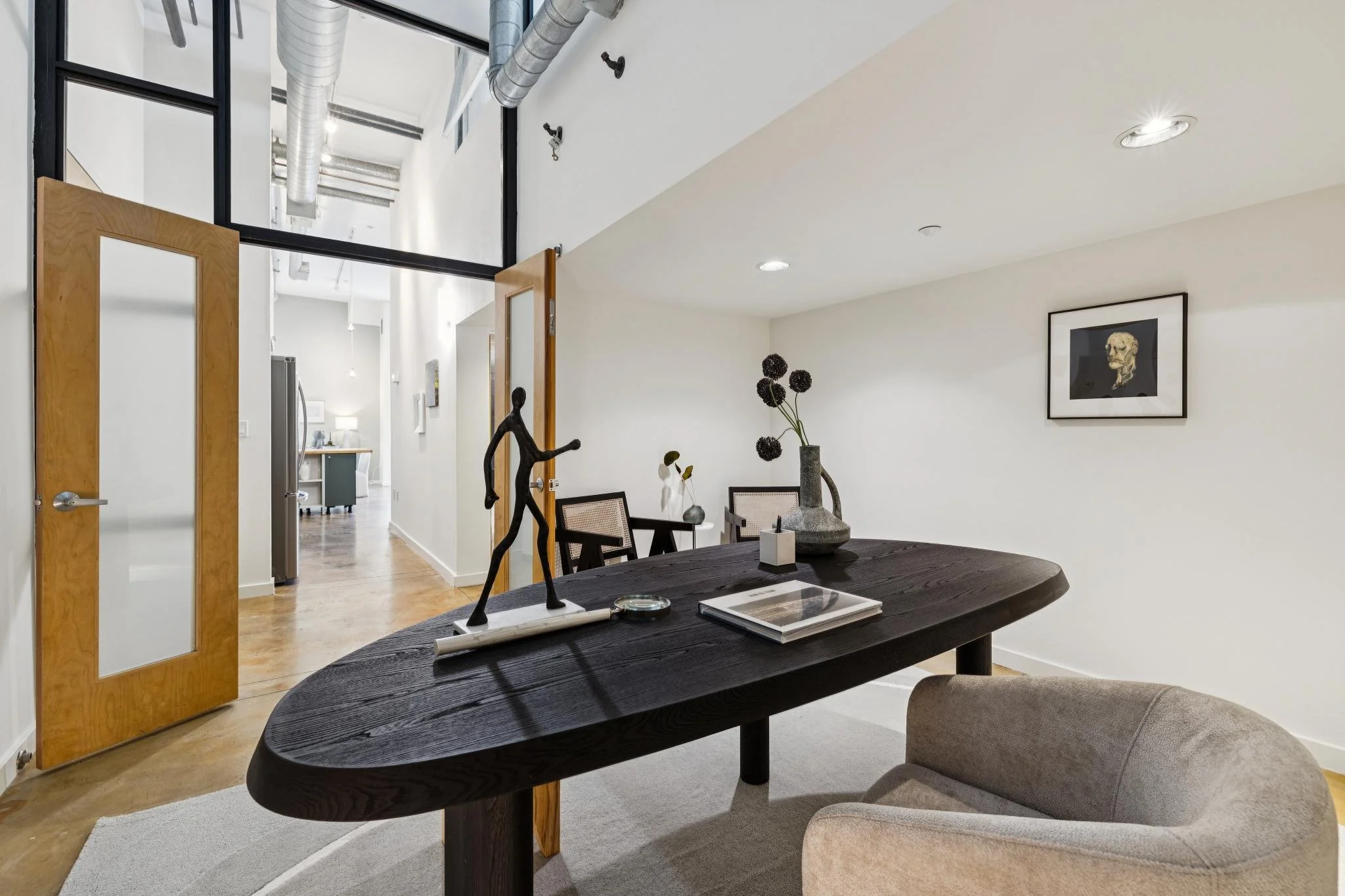 Interior of a modern apartment with a dining area featuring a dark wooden table, beige upholstered chairs, decorative sculpture, vase with flowers, and wall art. Background shows kitchen and hallway with wooden doors, white walls, and ceiling with du