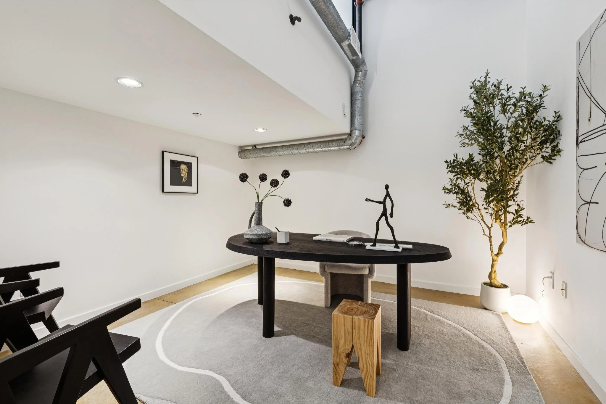 Modern interior of a room with a black round table, decorative sculptures, a tall potted plant, framed art on the wall, and a light-colored rug with circular patterns.