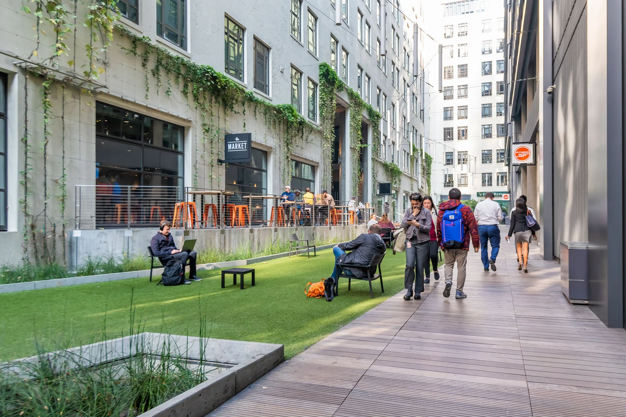 People walking and sitting in an urban outdoor space with grass, benches, and surrounding tall buildings, including a restaurant with an outdoor seating area.