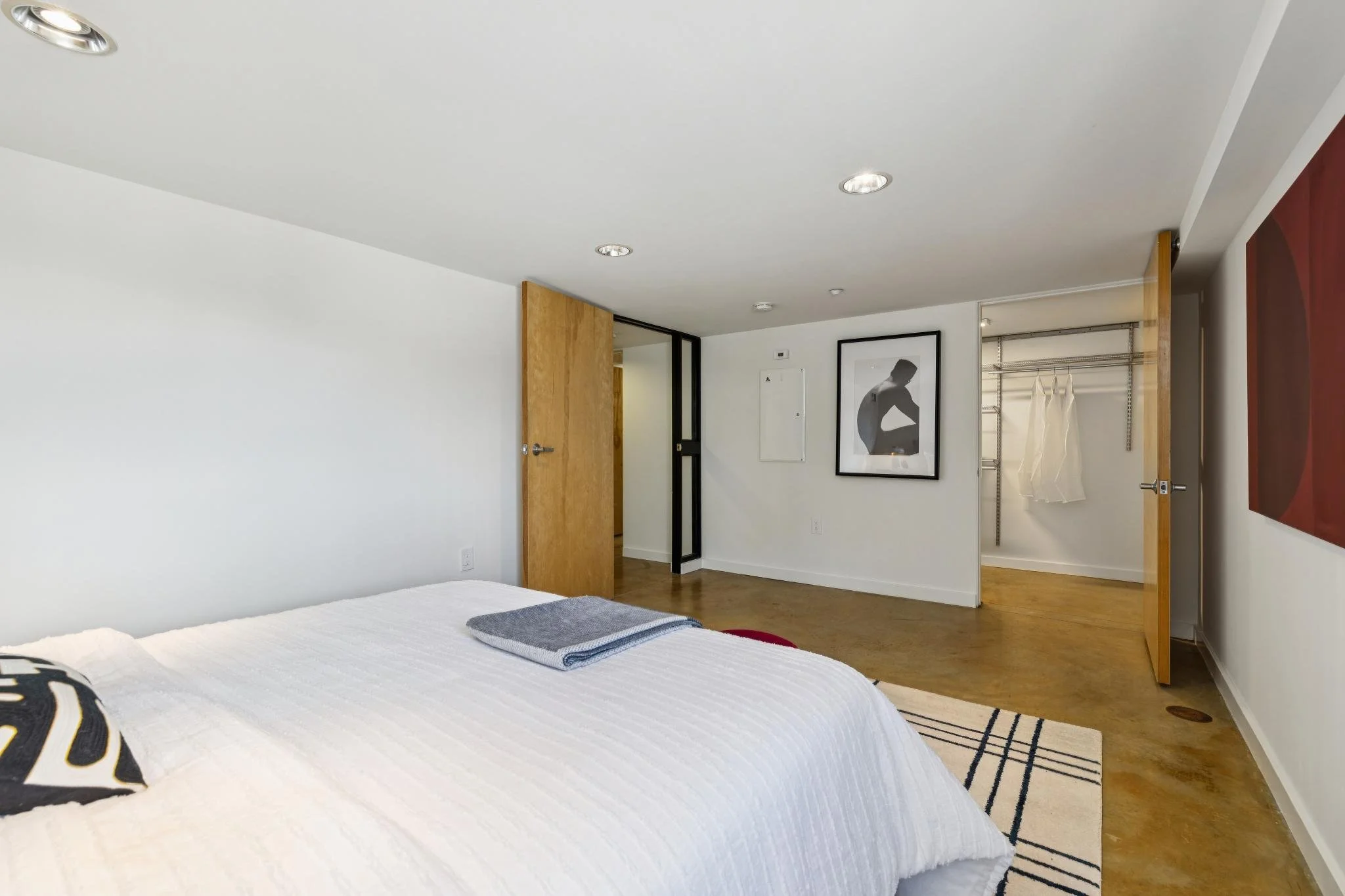 Modern bedroom with white walls, wooden doors, and framed black-and-white art. Part of a bed with white bedding and decorative pillows visible, along with a small striped rug.