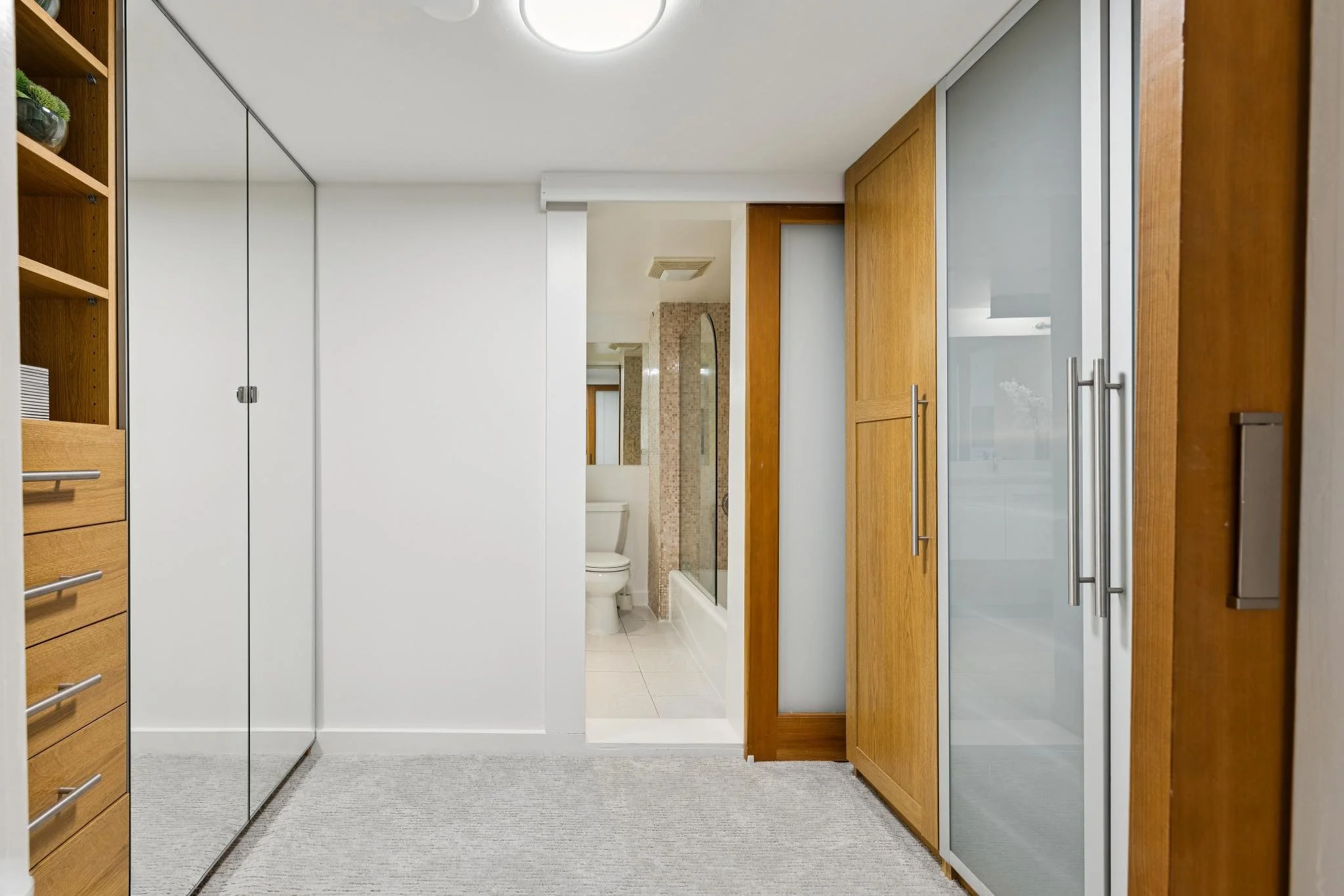 View of a modern bathroom with wood and glass cabinets, a white wall, a frosted glass sliding door, and a glimpse of a toilet in the background.