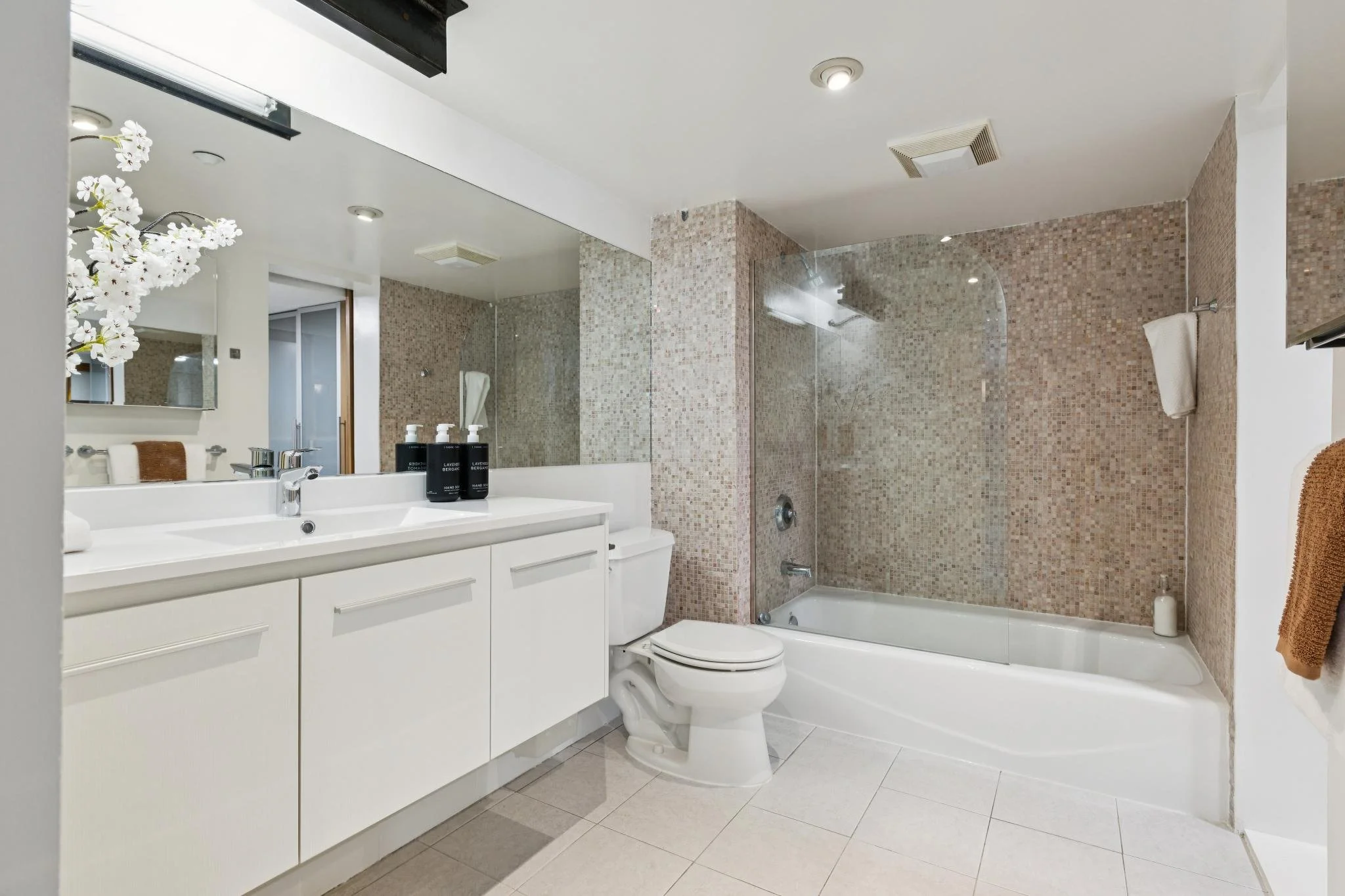 A modern bathroom with a white vanity, black soap dispensers, a mirror, a compact toilet, and a glass-enclosed shower with beige mosaic tiles. There is a folded towel on a hook and a small flower arrangement on the mirror's side.