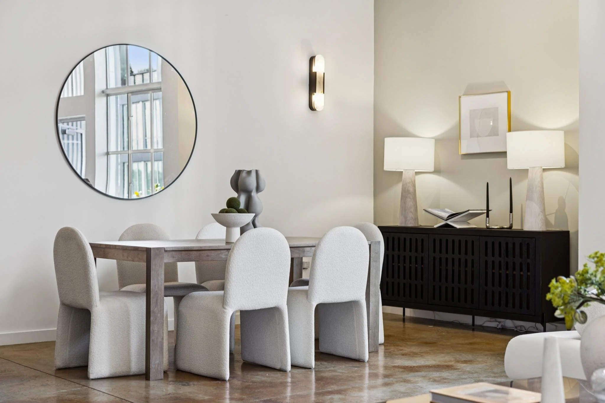 A modern dining room with six beige upholstered chairs around a wooden dining table. The table has a white ceramic vase with decorative spheres. There is a black sideboard with two tall beige table lamps, a picture frame, and a book on top. A large r